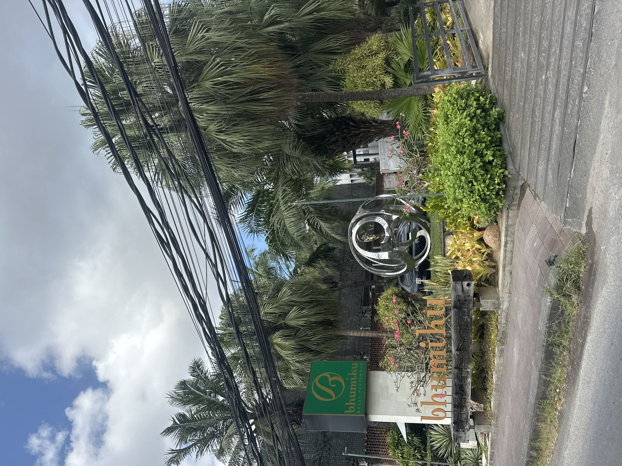 The image depicts the entrance to Bhumiku Balai Pertemuan, a venue in Denpasar, Indonesia. The scene is characterized by lush tropical vegetation, including several palm trees, and a prominent sign with the venue's name. A circular metal sculpture is positioned in front of the entrance, adding a modern artistic element to the tropical setting. Overhead, a dense network of electrical wires crisscrosses the sky, partially obscuring the view of the buildings and sky. The foreground shows a paved walkway and road, suggesting this is a street-level view. The overall atmosphere is one of a tropical, possibly humid, environment.