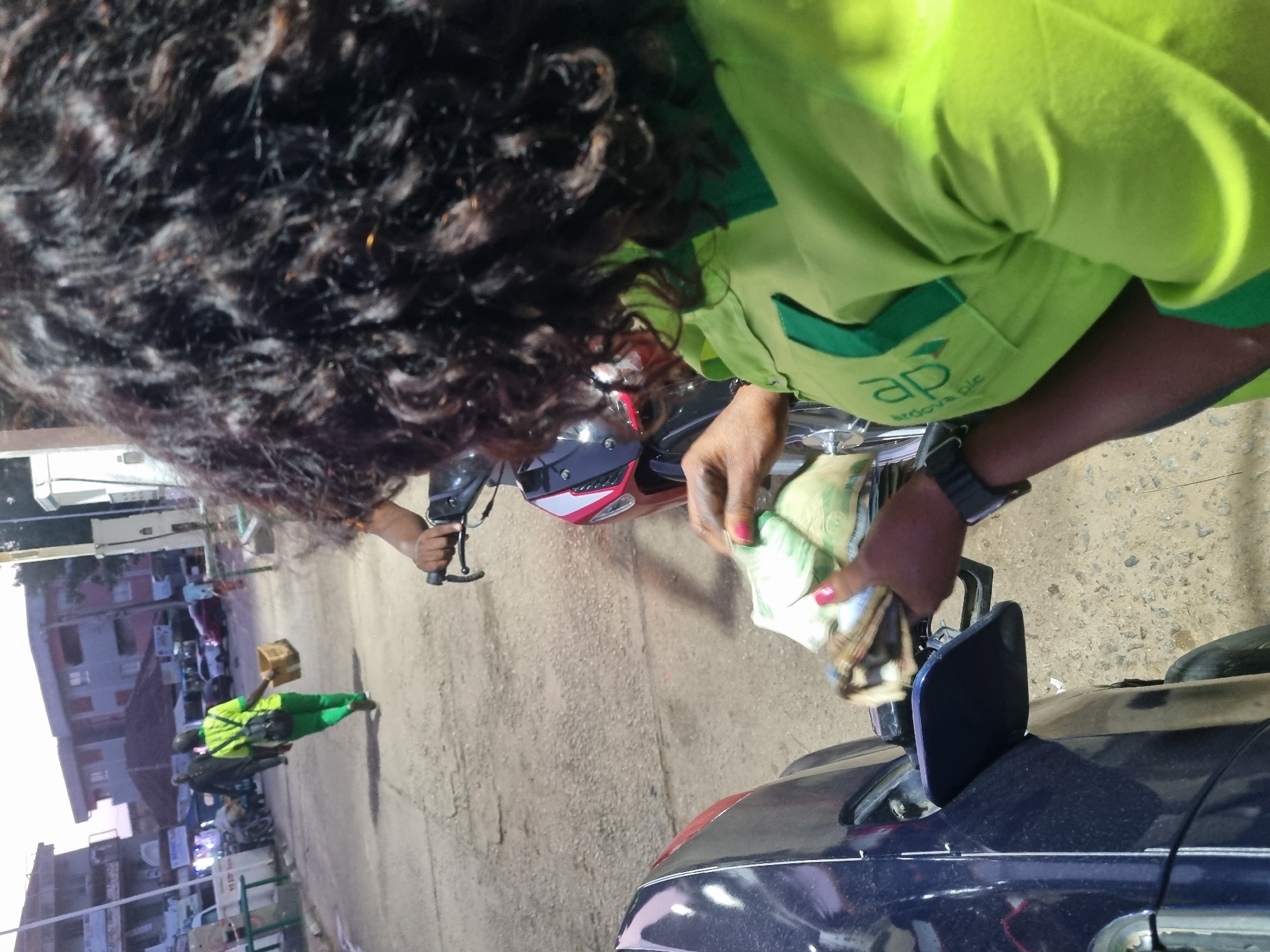A person wearing a light green uniform with the inscription "ap ardova plc" is counting money, likely Nigerian Naira, near the fuel tank of a dark blue car at what appears to be a filling station. A motorcyclist is visible in the background also wearing light green work clothes. The location seems to be Jos, Nigeria, based on contextual clues.