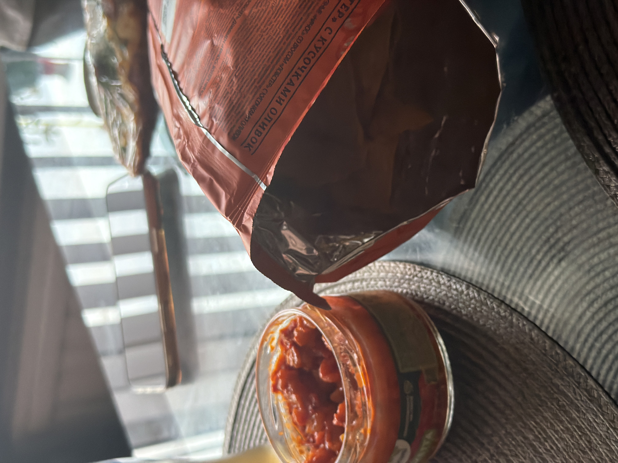 A close-up, low-angle shot captures a glass jar filled with a reddish relish or salsa, placed on a woven placemat. To the right, an open bag of red packaging, likely for snacks, is partially visible, with its contents obscured. The items are situated on a reflective surface, possibly a glass table, which shows reflections of striped blinds from a window in the background. A thin layer of amber liquid is visible in the jar, and the relish appears chunky with visible pieces of vegetables. The environment suggests an indoor setting, likely a dining area or kitchen. There is no indication of the city of Kolpino, Russia.