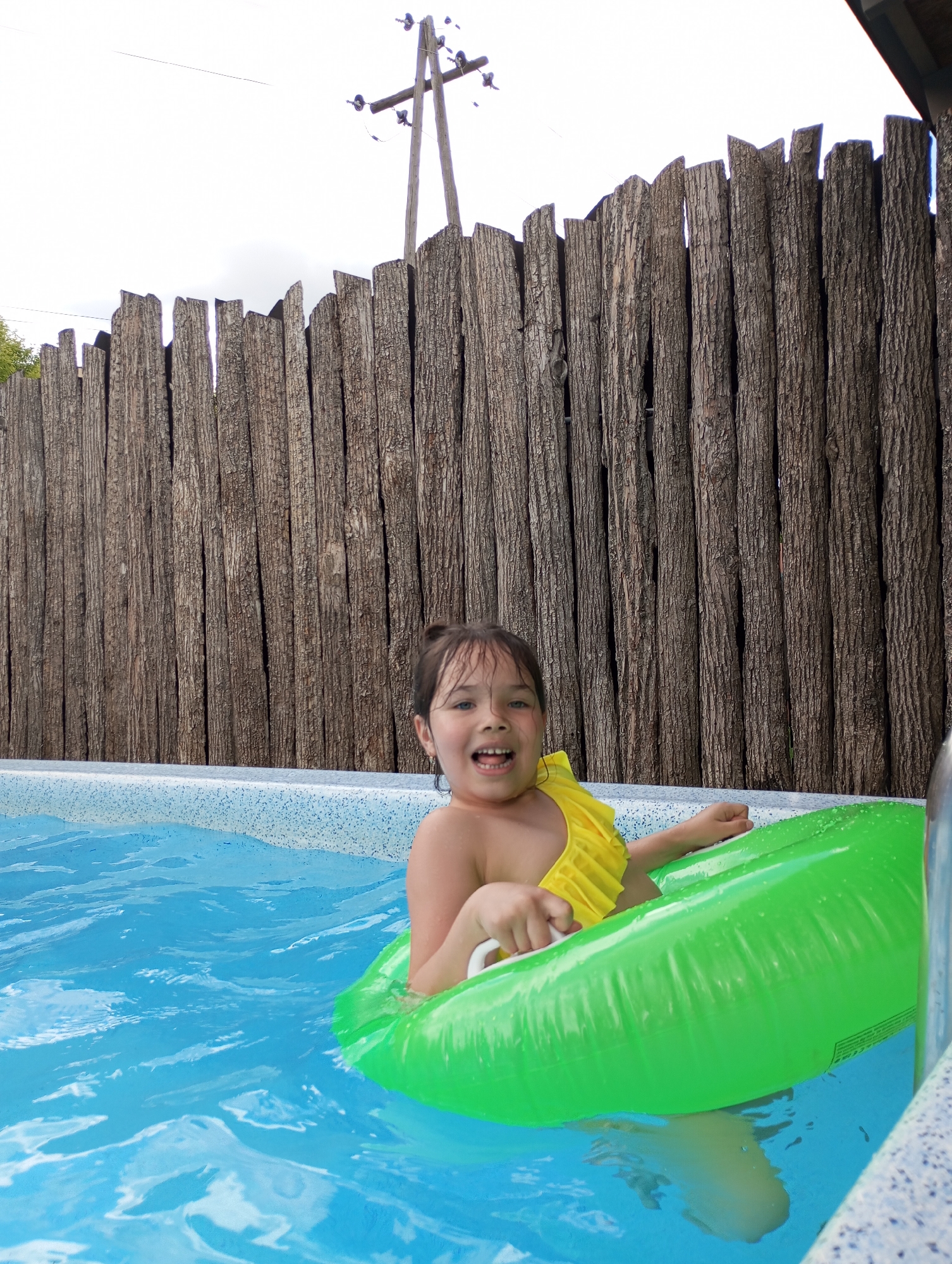 An outdoor, vertical shot depicts a young girl joyfully playing in a swimming pool. The girl, with dark hair and a wide, open-mouthed smile, looks directly at the camera, conveying happiness and excitement. She is wearing a bright yellow, ruffled swimsuit top and is partially submerged in the clear blue water, holding onto a vibrant green inflatable swim ring.
The pool edge features a light blue and white speckled pattern. Behind the pool and the girl, a tall, rustic fence made of rough-hewn vertical logs or planks forms a textured, dark brown backdrop. The fence appears weathered.
Above the fence, the sky is bright and largely overcast, indicating daytime conditions without direct harsh sunlight. To the upper right, a wooden utility pole with insulators and wires is visible against the bright sky. Hints of green foliage can be seen peeking over the fence on the left side of the frame. The scene captures a lively moment of leisure. No legible text is visible on any of the objects or surfaces in the image. The location is Izhevsk, Russia.