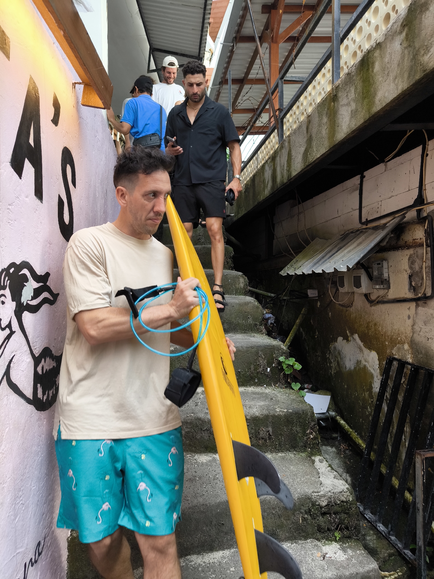 A man with short dark hair, wearing a tan t-shirt and turquoise shorts with a flamingo pattern, is holding a yellow surfboard with black fins. He is standing on concrete stairs leading down, and he appears to be looking to his left. On the wall to his left, there is graffiti artwork of a woman's face and the letter "A" in large black font. Behind the man, ascending the stairs, are three other men. One is wearing a black short-sleeved button-down shirt and black shorts, holding a phone, and wearing sandals. Behind him, one man is wearing a white t-shirt and a white baseball cap and the third man is wearing a blue shirt and a black baseball cap. The surroundings include a dilapidated building or structure next to the stairs, with exposed wires, metal roofing, and a black metal grate. The overall environment has a slightly unkempt or rundown appearance. The location is Uluwatu, Indonesia.