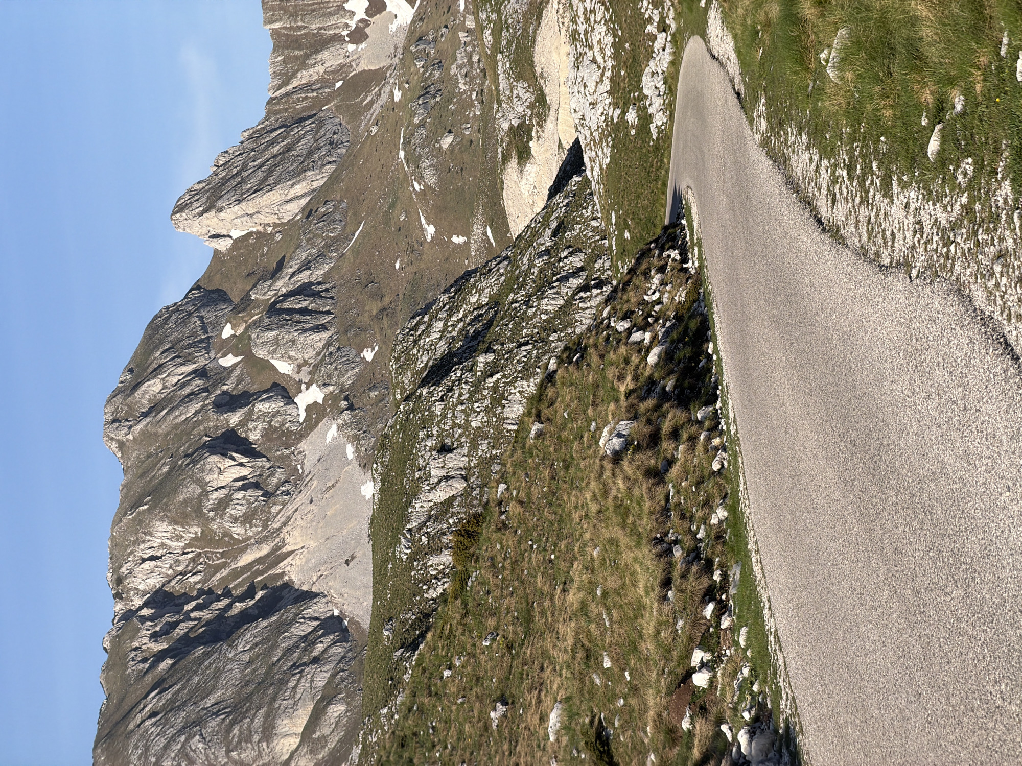 The image depicts a narrow asphalt road curving along the side of a mountain in Boričje, Montenegro. To the left of the road, a steep, rocky mountainside rises with patches of grass and lingering snow. The sky is visible in the upper left, a clear blue. The road surface is gray and appears to be well-maintained. To the right of the road is a sloping terrain covered with grass, rocks, and sparse vegetation. There are no people or animals visible in the frame. The photograph appears to have been taken during the day, with sufficient natural light.