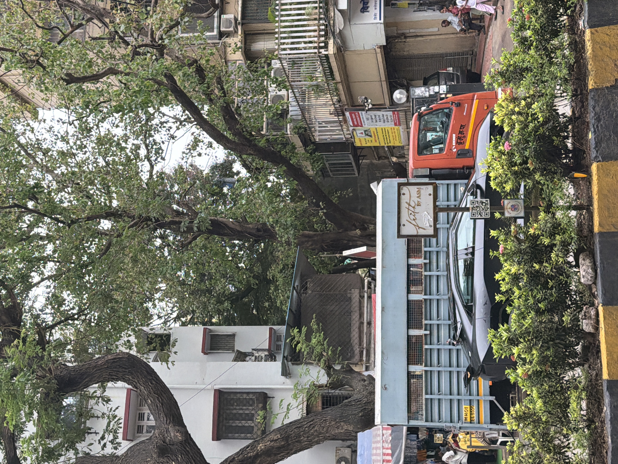 A street scene in Mumbai, India, captured on an overcast day. In the foreground, a black sports car with an aggressive rear spoiler is parked behind a planter box filled with green foliage and some small pink flowers. The car's license plate reads "MH01 EE3556". To the left of the car, a light blue truck with a metal cargo bed is partially visible. A sign on the truck's side reads "faith BY ASM". To the right of the truck is a bright orange EICHER truck, with text in Hindi on its side. Further back, a multi-story building with white and beige facades features several windows and air conditioning units. A white metal railing adorns a balcony on one of the upper floors. A large tree with thick branches and green leaves dominates the upper portion of the frame, its branches extending across the buildings and sky. In the background, people are seen sitting on a bench outside a shop. The overall impression is a bustling urban environment with a mix of vehicles, architecture, and natural elements.