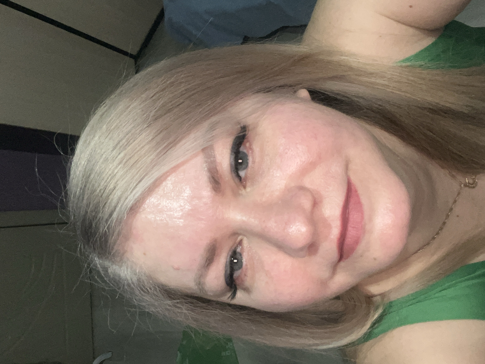 A medium shot of a fair-skinned person with blonde hair, light-colored eyes, and pink lips, wearing a green top. A gold chain necklace is visible on their neck. The background appears to be an indoor space with light-colored walls and a dark-colored panel in the upper left corner. There is no verifiable evidence to support any claims regarding a location in Izhevsk, Russia.