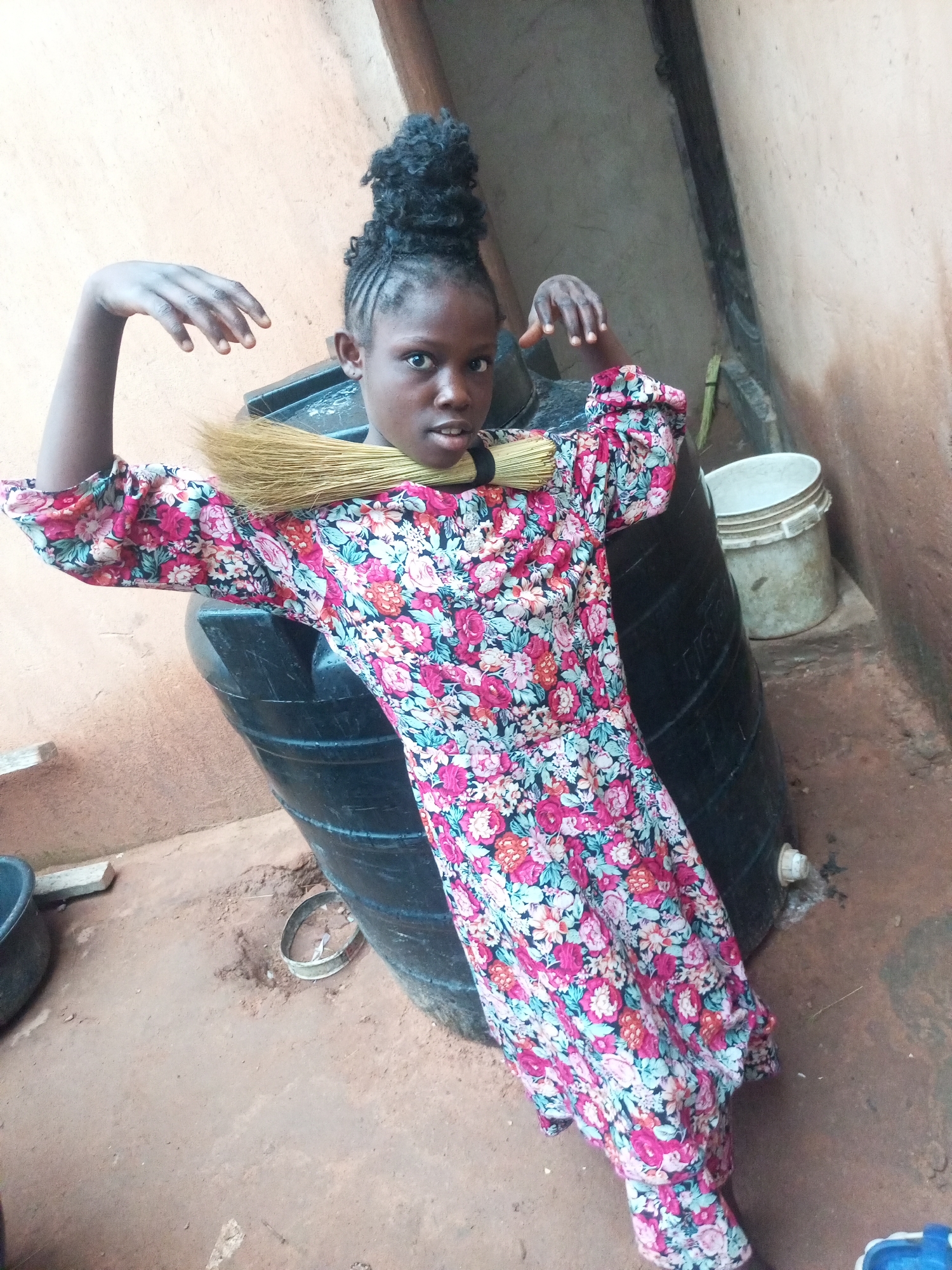 In Tumfure, Nigeria, a young Black girl with her hair styled in braids pulled up into a bun stands in front of a large black water tank. She is wearing a colorful floral dress with pink, white, and blue flowers. A broom is draped across her shoulders. Her arms are raised, and her hands are spread open. To her right, a white plastic bucket sits on the ground. The ground is a mix of dirt and pebbles, and a rough-textured wall forms the background. The scene appears to be in an outdoor or semi-outdoor setting, possibly a yard or an alleyway.