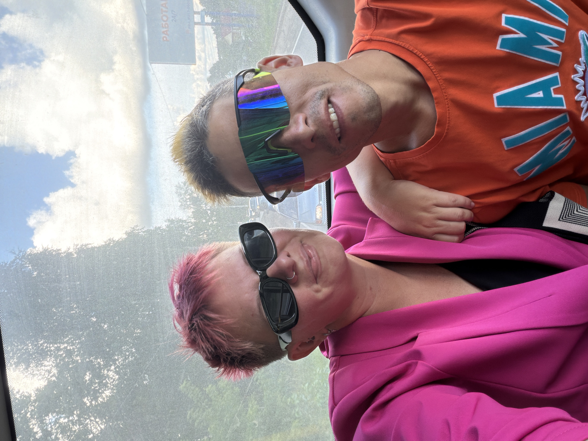 Two individuals are captured in a selfie, likely within a vehicle, with a bright, slightly overexposed exterior view visible through a window. The person on the left, with short, pink-tinted hair and dark sunglasses, wears a vibrant pink blazer over a black top. The person on the right, with spiky, grey-streaked hair and distinctive iridescent wraparound sunglasses, sports an orange t-shirt with the word "MIAMI" printed in teal and white. The background suggests an outdoor setting with trees, a road, and a billboard that displays Russian text and the number "24/7," indicating a service available around the clock. The overall impression is a candid moment, possibly during a road trip, with the individuals appearing relaxed and engaged with the camera. The location is identified as Chaikovsky, Russia.