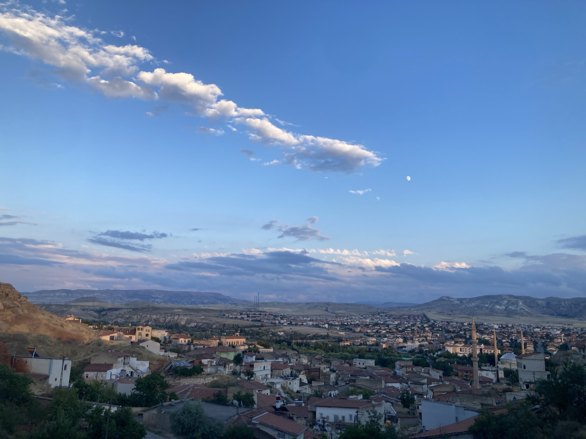 This is a wide-angle, high-angle shot of Avanos İlçe Merkezi, Turkey, taken during daylight hours. The sky is a bright blue with scattered white clouds, and a sliver of the moon is visible in the upper right quadrant. The foreground is dominated by a hillside with scattered trees and houses. The houses are mostly made of stone and have red tile roofs.
In the mid-ground, a town spreads out, characterized by a dense collection of buildings with red tile roofs. Several minarets rise above the rooftops, indicating the presence of mosques. The town is nestled within a valley, with rolling hills and mountains forming the backdrop. The landscape appears arid and rocky, with sparse vegetation. The overall impression is of a traditional Turkish town set against a natural landscape. There are no people visible in the image.