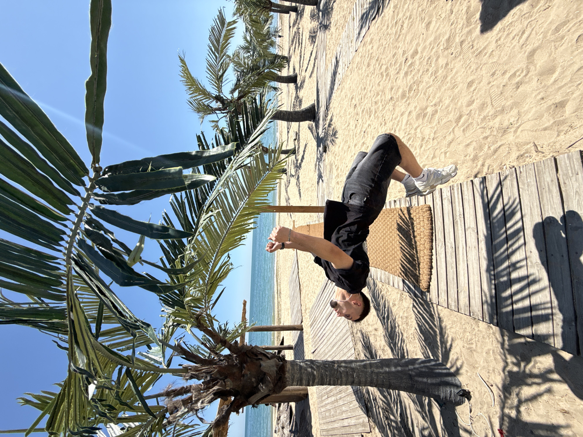 A man dressed in black shorts and a black shirt, wearing white shoes, is doing a handstand on a wooden boardwalk near a beach. Palm trees surround him. The location is identified as Varna, Bulgaria, based on the beach environment. Sand is visible alongside the boardwalk. The sky is clear and blue.