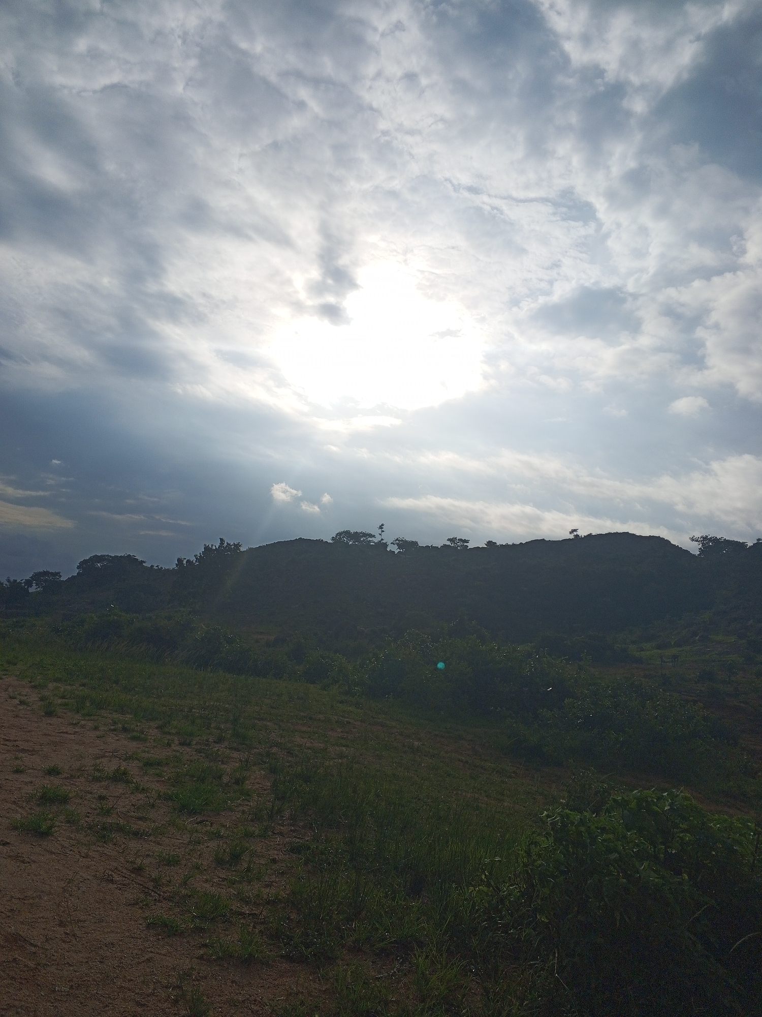 The image depicts a landscape scene with a bright sky filled with scattered clouds. The sun is prominently visible, casting rays of light through the cloud cover. Below the sky is a silhouetted hill covered in dark green vegetation, with trees dotting the crest. In the foreground, there is a patch of dry, sandy ground with sparse green grass. The overall impression is of a natural, undeveloped area. The location is identified as Gwong, Nigeria, suggesting this is likely a view of the natural surroundings in or near the city. There are no people or specific objects of human construction visible in the image. The environment appears to be rural or semi-rural, with rolling terrain and abundant greenery under a dynamic sky.