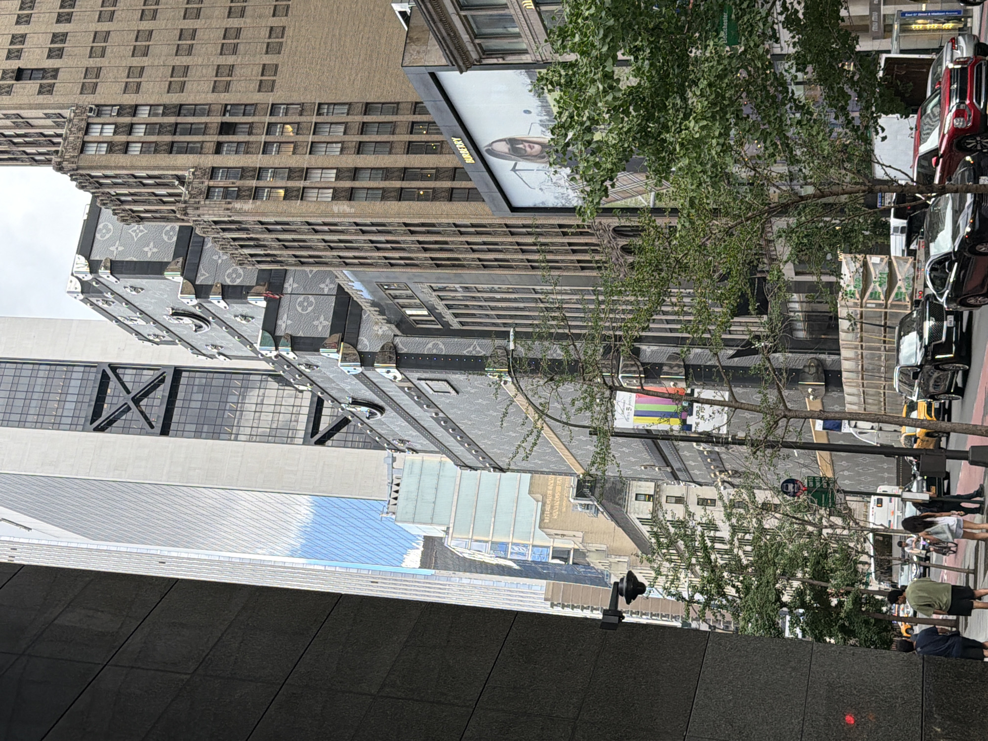 This is a street-level photograph taken in New York City, United States of America, depicting a commercial street with several tall buildings. The perspective is from sidewalk level, looking up at the architecture and street activity. Dominating the foreground on the left is a dark, textured building facade, partially obscuring a view further down the street.
On the right side of the street, a prominent Louis Vuitton flagship store is visible, adorned with oversized, iconic LV trunk motifs. This store spans several stories and features metallic accents and detailing. Adjacent to it, a large billboard displays an advertisement for Burberry, featuring a woman wearing sunglasses and plaid clothing. Further down the street, other buildings with various architectural styles and window patterns are visible, including a tall, modern skyscraper with reflective glass and a more traditional brick building with numerous windows.
The street itself is busy with traffic. Several cars are present, including a black SUV, a black sedan, a red SUV, and yellow taxis. Pedestrians are also visible on the sidewalk, including a group of three individuals on the left and a couple on the right. One person is riding a bicycle further down the street. Green trees line the sidewalk, providing some natural element amidst the urban landscape. Traffic signs and streetlights are also present. The sky appears overcast. The overall impression is a bustling urban commercial district in New York City.