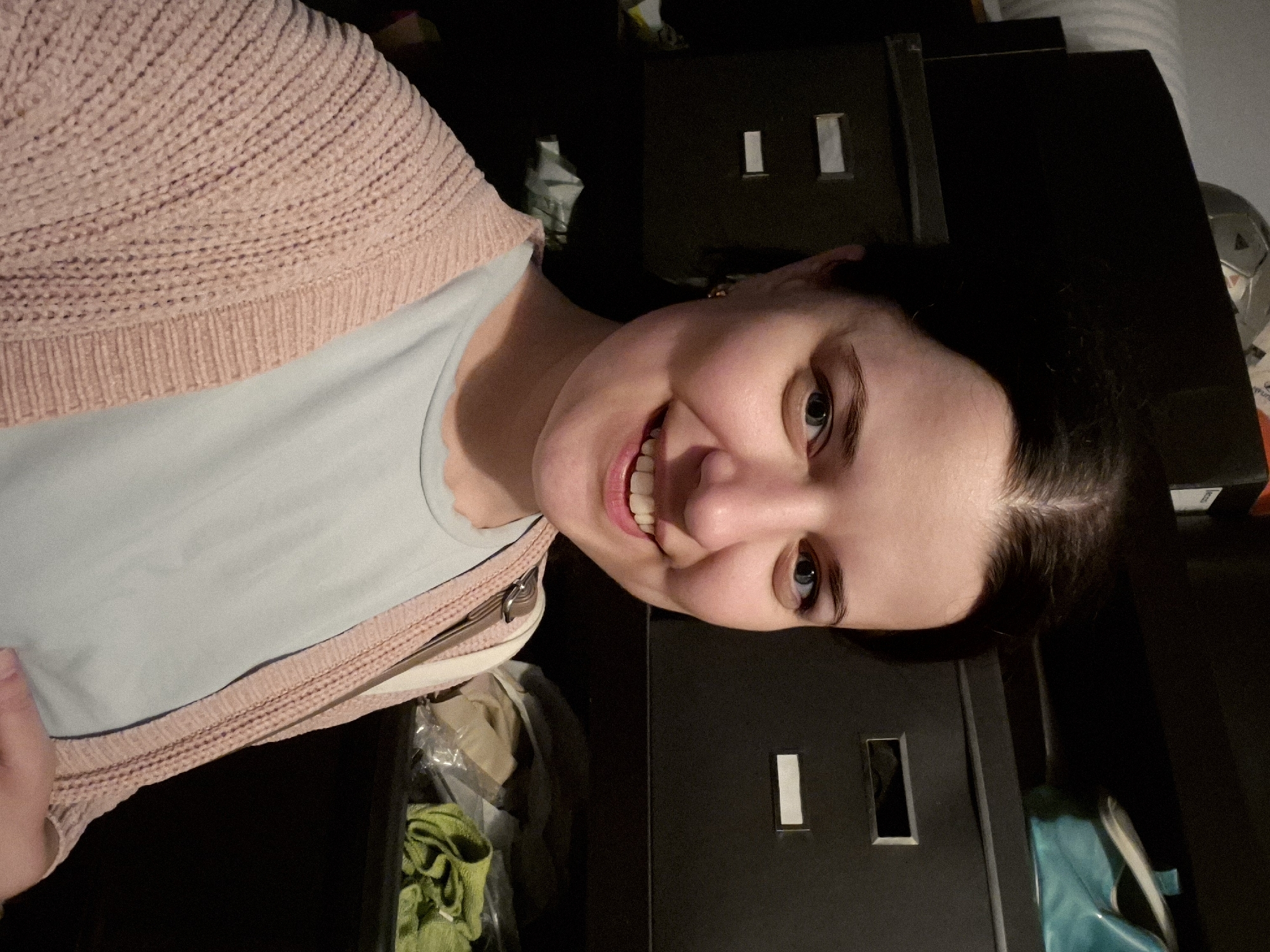 A woman is taking a selfie in front of dark-colored shelves. She has dark hair, blue eyes, and is wearing a light blue shirt and a pink cardigan. Her mouth is open in a smile, and her teeth are visible. Behind her, the shelves contain various items, including what appear to be fabric bins and a teal bag. The lighting in the image is dim, casting shadows. The background appears to be an indoor domestic setting, such as a closet or storage area. The image does not contain information that allows for its location in Saint Petersburg, Russia.