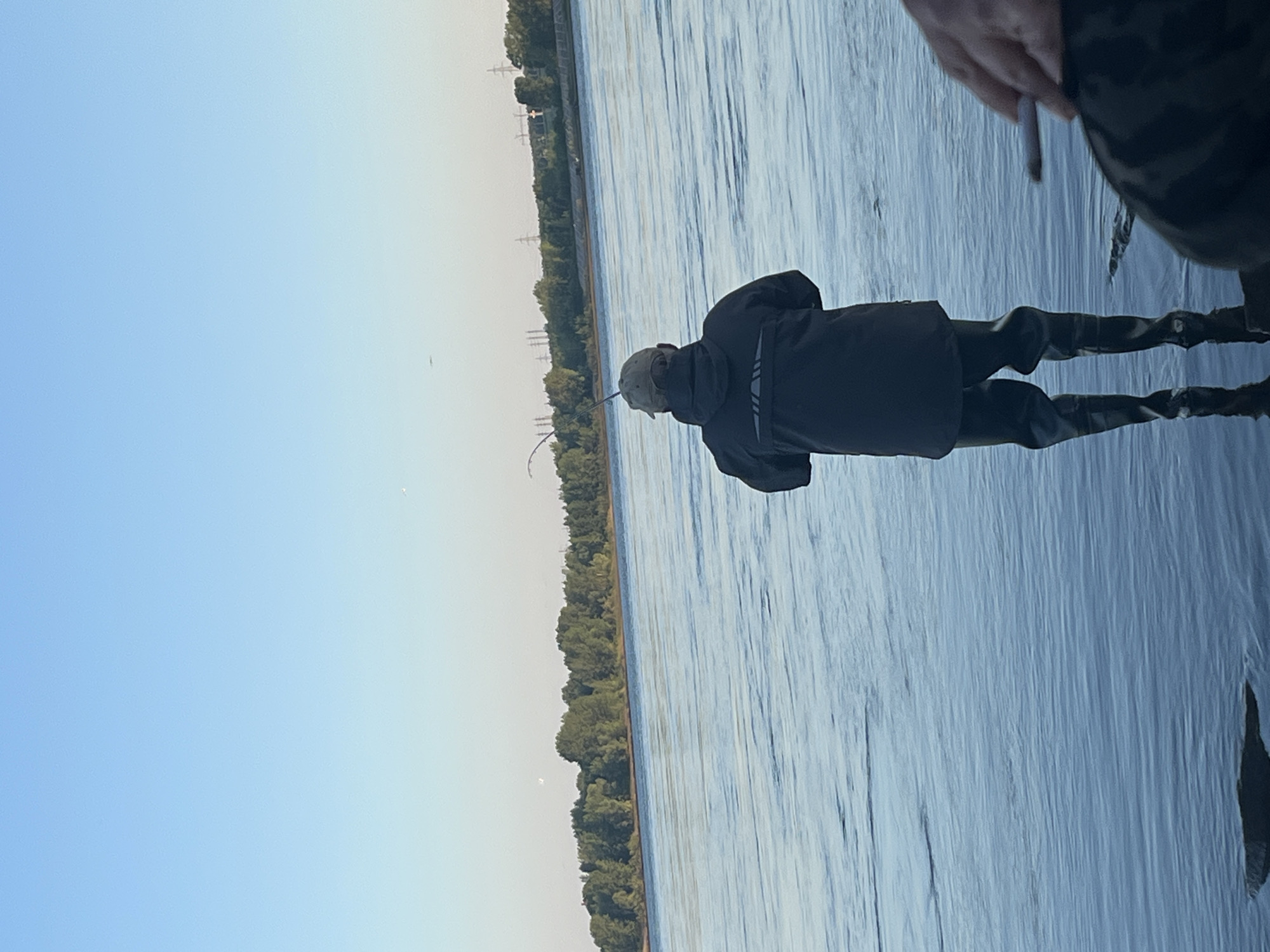 A man wearing a dark jacket and waders stands on the shore of a body of water, fishing. He is facing away from the camera, and holding a fishing rod. The water is calm and reflects the blue sky and green trees on the opposite bank. In the distance, there are power lines and industrial structures. The image appears to be taken in Chaikovsky, Russia, given the context of other images from the same location. Another person's hand, holding what appears to be a cigarette, is visible in the lower right corner of the image.