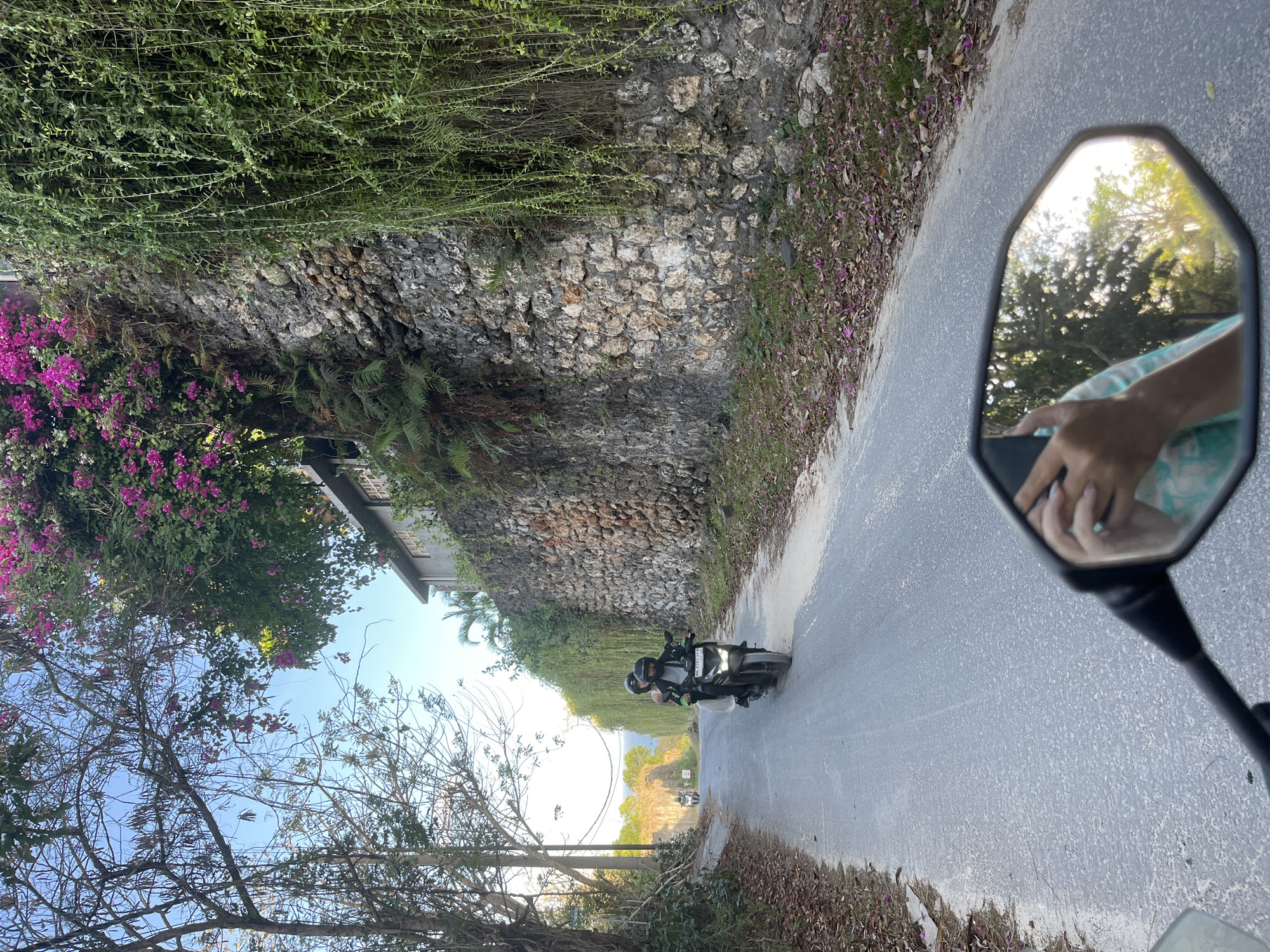 A motorcycle with two occupants, wearing helmets, travels on a paved road in Kuta Selatan, Indonesia. The road is lined with trees on the left and a stone wall covered with vines and flowering plants on the right. The motorcycle is being ridden by a person wearing a black jacket, and a second person is seated behind them. In the foreground, a motorcycle's rearview mirror reflects the rider's hand holding a phone and the surrounding environment. The sky is clear and blue, suggesting a sunny day. The area appears to be a rural or semi-rural setting, with lush vegetation and a natural stone wall.
