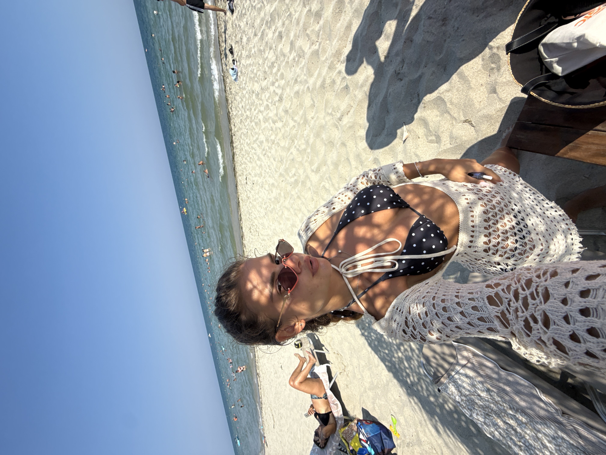 A woman is taking a selfie on a beach in the city of Katerini Municipality, Greece. She is wearing a black and white polka dot bikini, a white crochet cover-up, and sunglasses. She has her hair in a braid. In the background, other people are swimming in the ocean and lounging on the sand. There are beach chairs and bags visible. The sky is clear and blue, and the sun is shining.