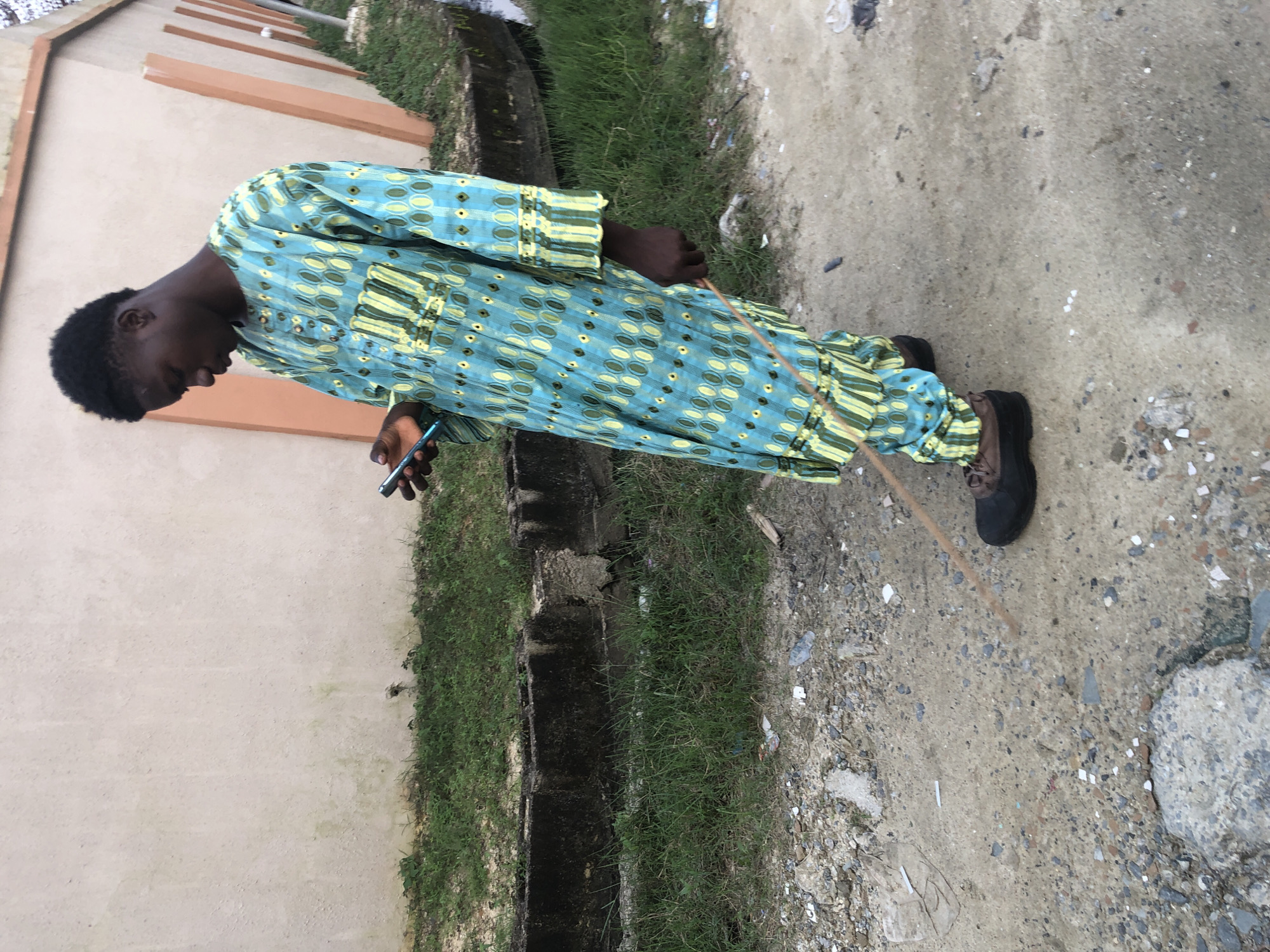 In Ado, Nigeria, a young Black male stands on a dirt path, looking down at a smartphone in his right hand. He is wearing a traditional blue and green patterned garment with yellow accents, long sleeves, and matching trousers. He is also wearing dark shoes with brown laces. In his left hand, he holds a thin, light-brown stick. The background consists of a light-colored building with orange accents, a strip of green grass, and a concrete drainage channel. The ground is a mix of dirt, small stones, and scattered debris. The lighting suggests it is daytime, possibly overcast.