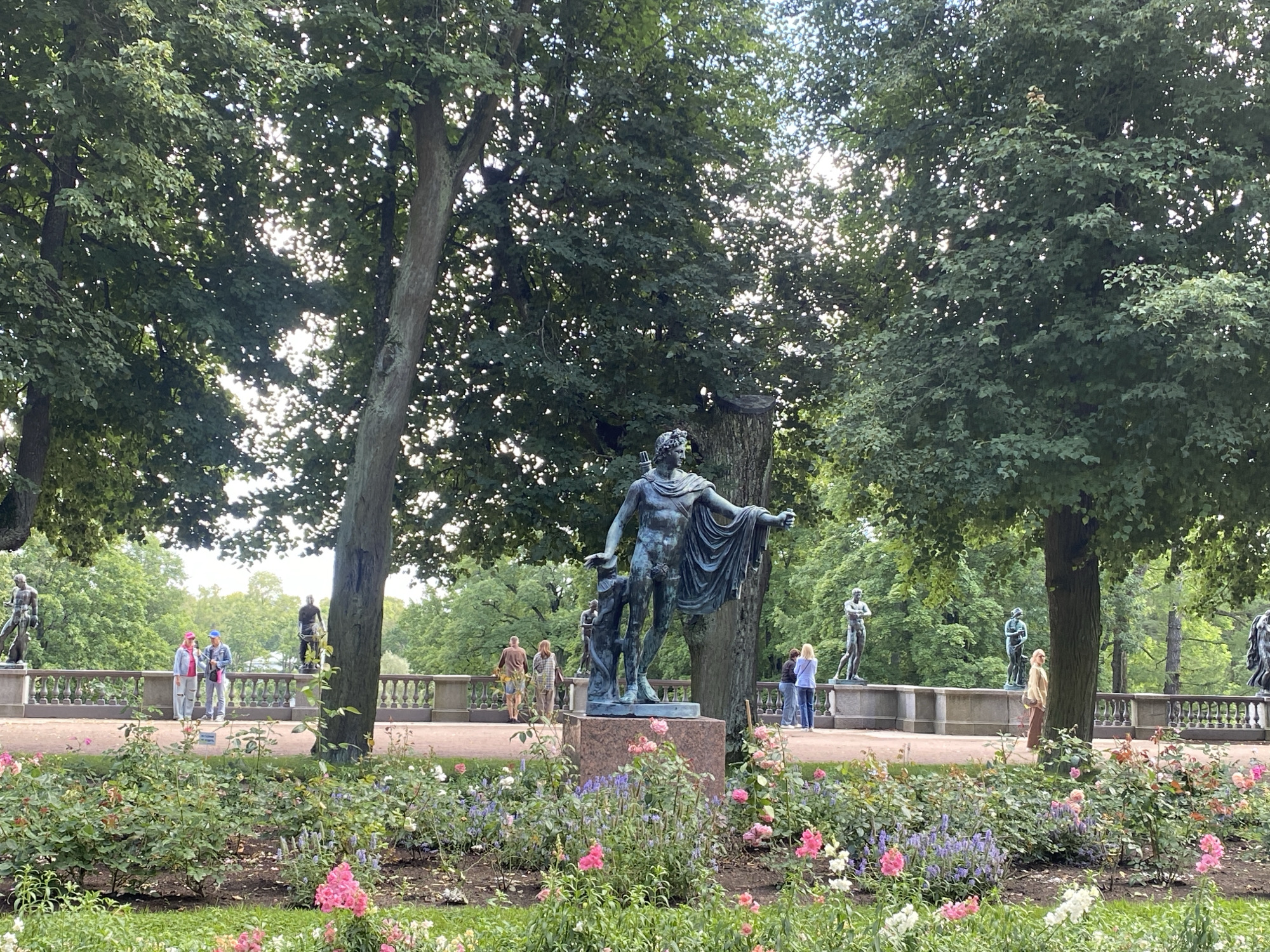 The image depicts a scenic outdoor setting in Pavlovsk, Russia, featuring a park with classical sculptures and lush floral arrangements. In the foreground, a vibrant flowerbed showcases a variety of blooming plants, predominantly pink roses and blue-purple hydrangeas, set against a backdrop of green grass. A large, dark bronze statue of a male figure, possibly a god or mythological hero, stands prominently on a stone pedestal in the mid-ground, with another smaller statue of a child at its side. Behind this central sculpture, a decorative stone balustrade runs along a pathway. Several other classical statues are visible along this balustrade, spaced apart and placed on pedestals.
A few individuals are scattered throughout the park, observing the sculptures and enjoying the environment. On the left side of the balustrade, two people, one wearing a pink hat and the other a blue hat, are standing and facing left. Further along the balustrade, near the center, two more individuals are walking away from the camera. To the right of the central statue, a person in a light-colored jacket and trousers is walking towards the right. On the far right side of the balustrade, another individual is visible, looking towards the sculptures.
The park is characterized by mature trees with dense green foliage, filtering sunlight to create dappled patterns on the ground. The overall atmosphere is serene and picturesque, suggesting a popular recreational area within the city of Pavlovsk.