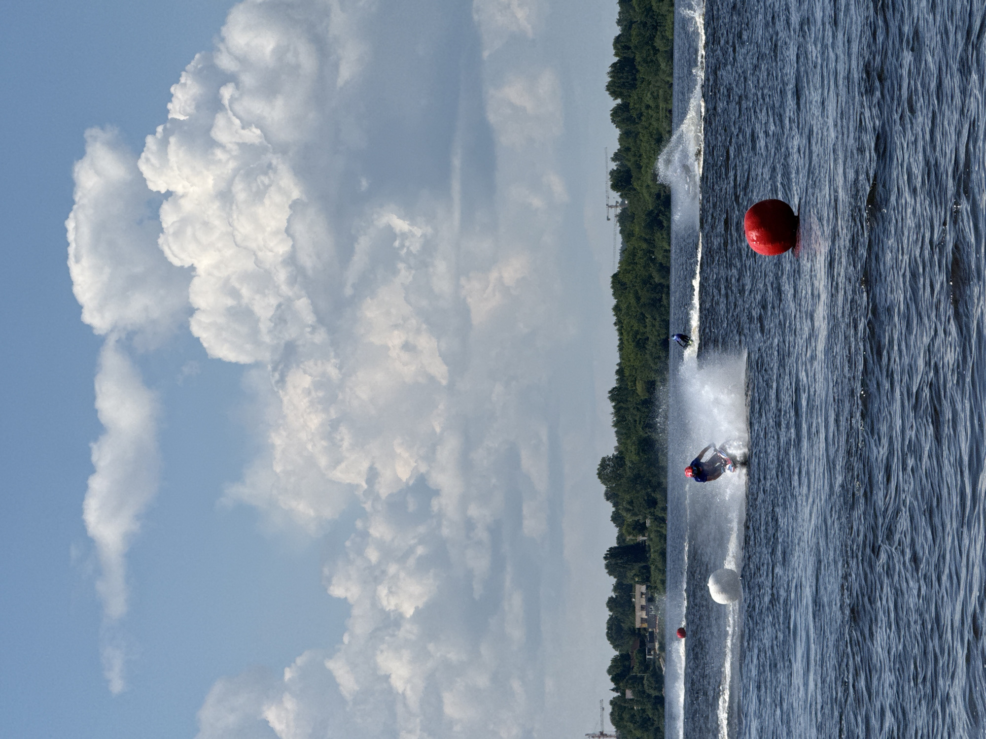 An action shot captures a jet ski race on a wide, dark blue body of water. The sky above is a bright blue, dotted with large, fluffy white clouds. In the foreground, a red buoy floats on the surface. A jet ski with a rider wearing a red helmet and blue top is in motion, creating a spray of water behind it. Further in the distance, another jet ski is visible, also generating a water spray. A white buoy is also present in the mid-ground. The background features a dense line of green trees and some buildings, with a construction crane visible in the far distance. This scene is likely taking place on the Kama River in Perm, Russia, given the presence of the river and the distinctive industrial elements in the background.