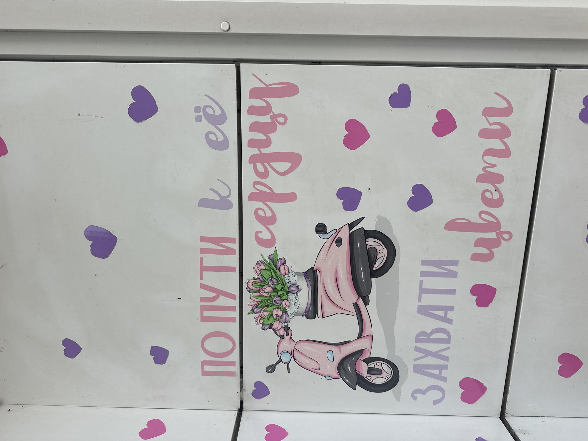 The image shows a white metallic surface, possibly a locker or a cabinet, with painted pink and purple decorations. The decorations include several hearts of varying sizes and two main phrases written in stylized Cyrillic script. The top phrase reads "ПО ПУТИ К ЕЁ" in pink and purple. Below this, a pink cartoon scooter with a basket of tulips is depicted. The bottom phrase reads "ЗАХВАТИ ЦВЕТЫ" also in pink and purple. The overall aesthetic is whimsical and cheerful, with a clear theme related to flowers and romance. The image was taken in Lytkarino, Russia.