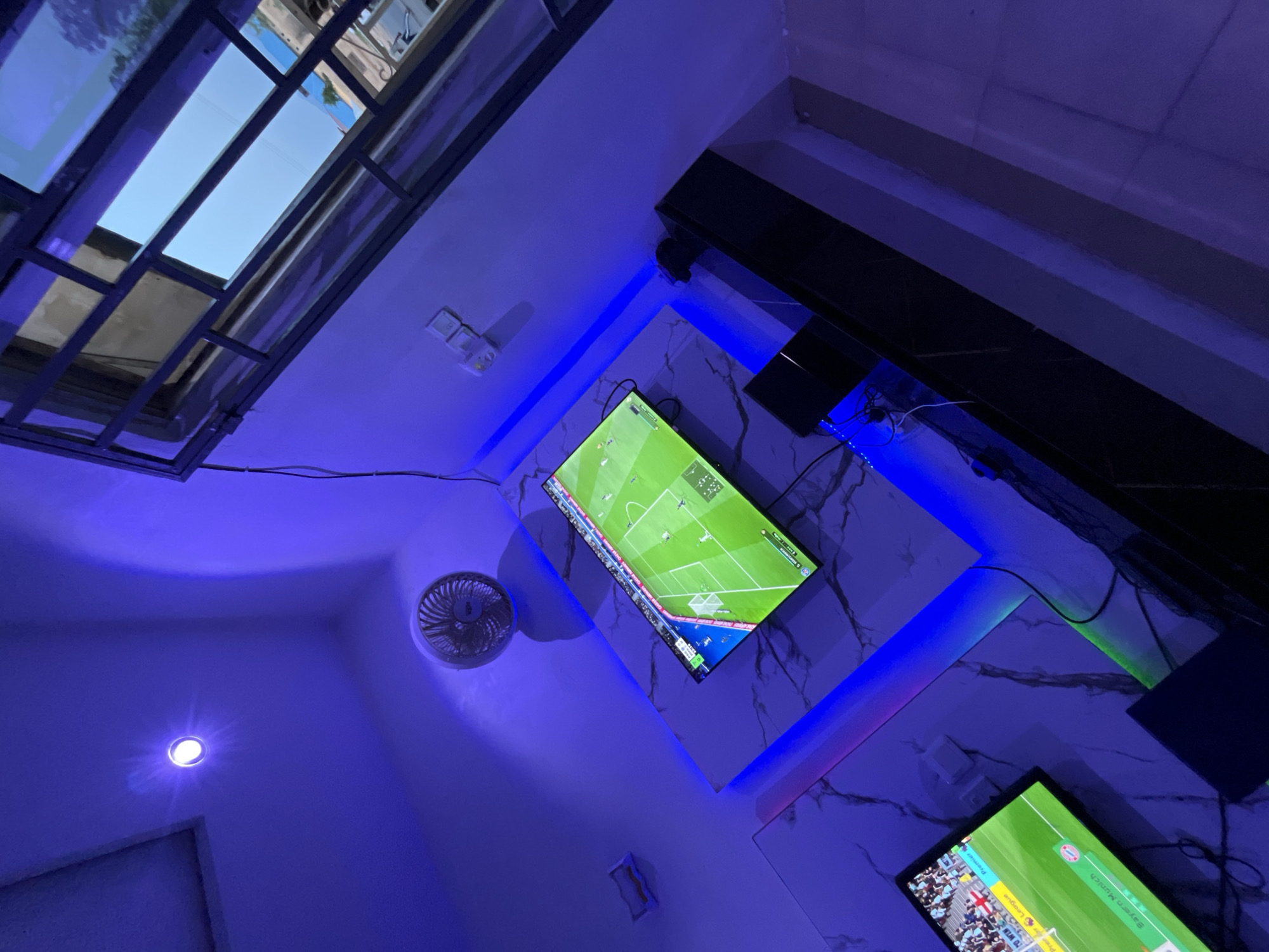 The image depicts an indoor scene illuminated by blue and purple LED lights. Two flat-screen televisions are visible, both displaying what appears to be a soccer video game. One television shows a game with players in white uniforms on a green field, and the other displays a game with teams labeled "Premier League" and "Bayern Munich." The televisions are mounted on a wall with a marble-like pattern. There is a small circular fan mounted on the ceiling. A window with metal bars is visible in the upper right portion of the image, looking out onto an outdoor environment. The overall impression is of a modern entertainment setup, likely in a residential or commercial space in Jalingo, Nigeria.