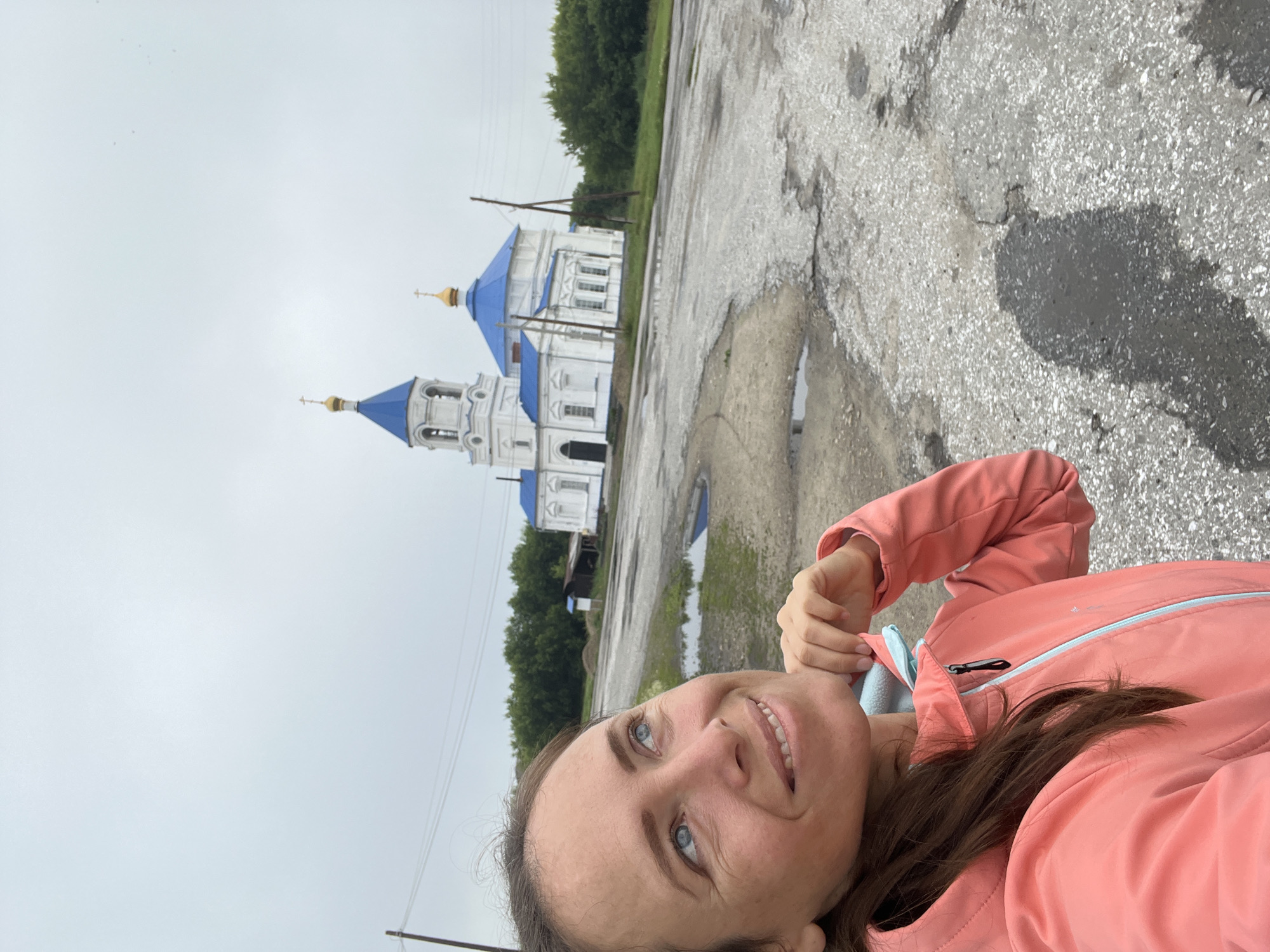A woman in a coral-colored jacket is shown in a close-up, selfie-style shot, looking up and to the right with a slight smile. Her right hand is raised to her chin, and her long brown hair is pulled back. In the background, a white Orthodox church with a blue roof and a bell tower stands under a cloudy sky. The church is situated on the outskirts of Pivkino, Russia, in an area with a damaged asphalt road, scattered puddles, and sparse vegetation. Power lines and a wooden building are also visible.