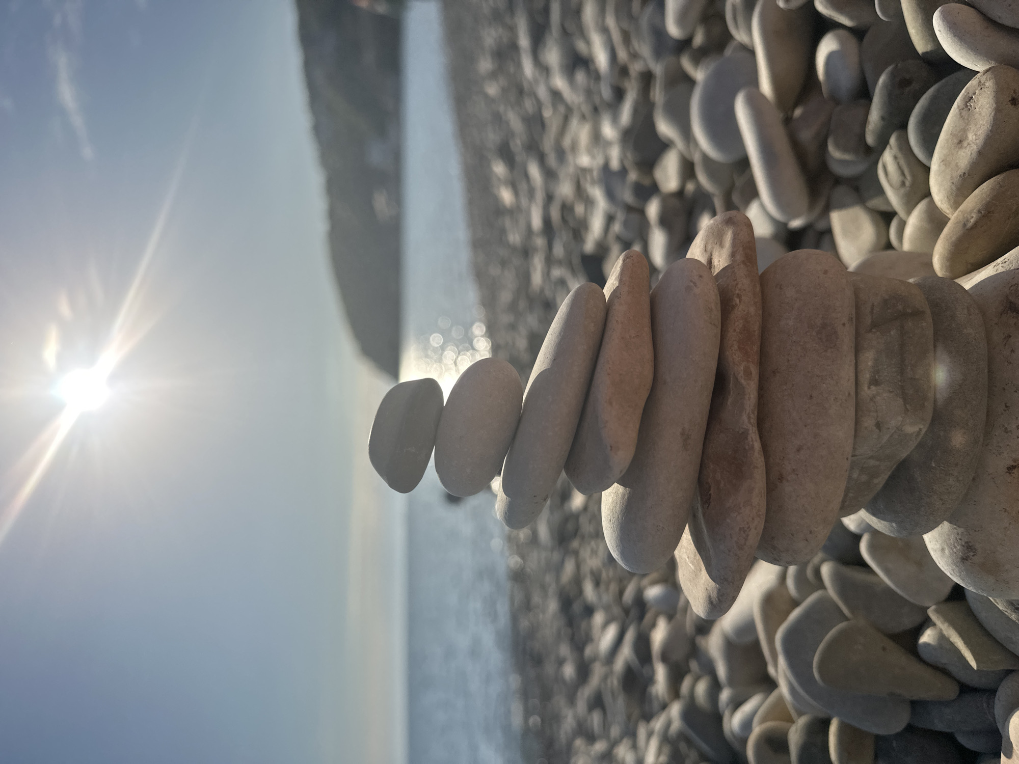 The image shows a stack of smooth, gray stones balanced on a pebble beach in Novomikhaylovsky, Russia. The stack consists of about a dozen stones, aligned in a row, creating an abstract sculpture. The background features the sea, the sky with the sun, and a faint outline of the coastline. Other scattered stones cover the beach surface. The lighting suggests a sunny day.