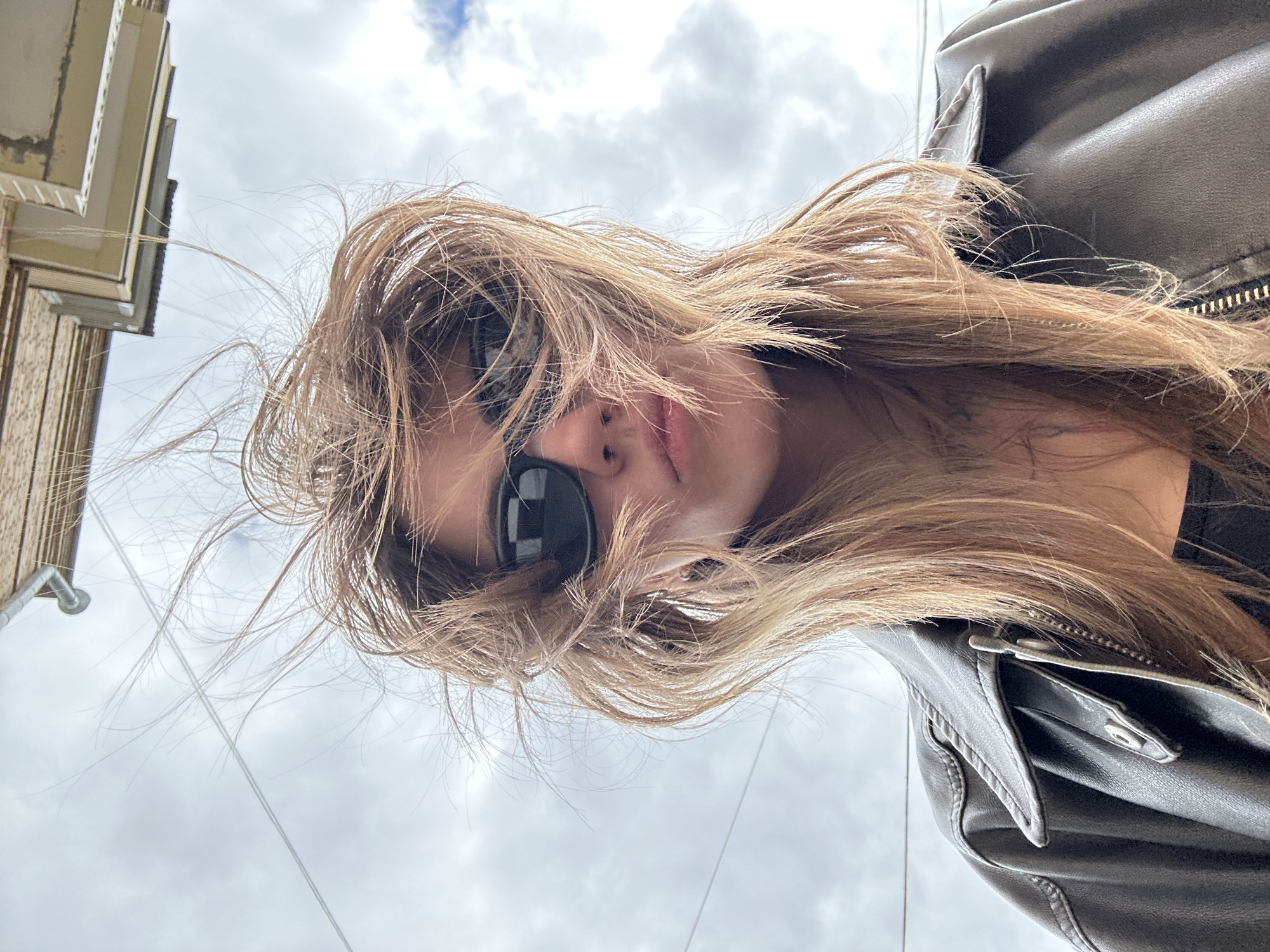 The image is a close-up, low-angle selfie of a young woman with light brown hair blowing in the wind. She is wearing dark sunglasses and a dark leather jacket with a zipper. The background is an overcast sky with clouds, and a building overhang is visible at the top of the frame. There are also thin wires or cables visible against the sky, suggesting an urban environment. The angle and composition indicate the photo was taken from below, looking up at the woman. The location is not specified in the image, and no elements are present that would definitively place it in Moscow, Russia.
