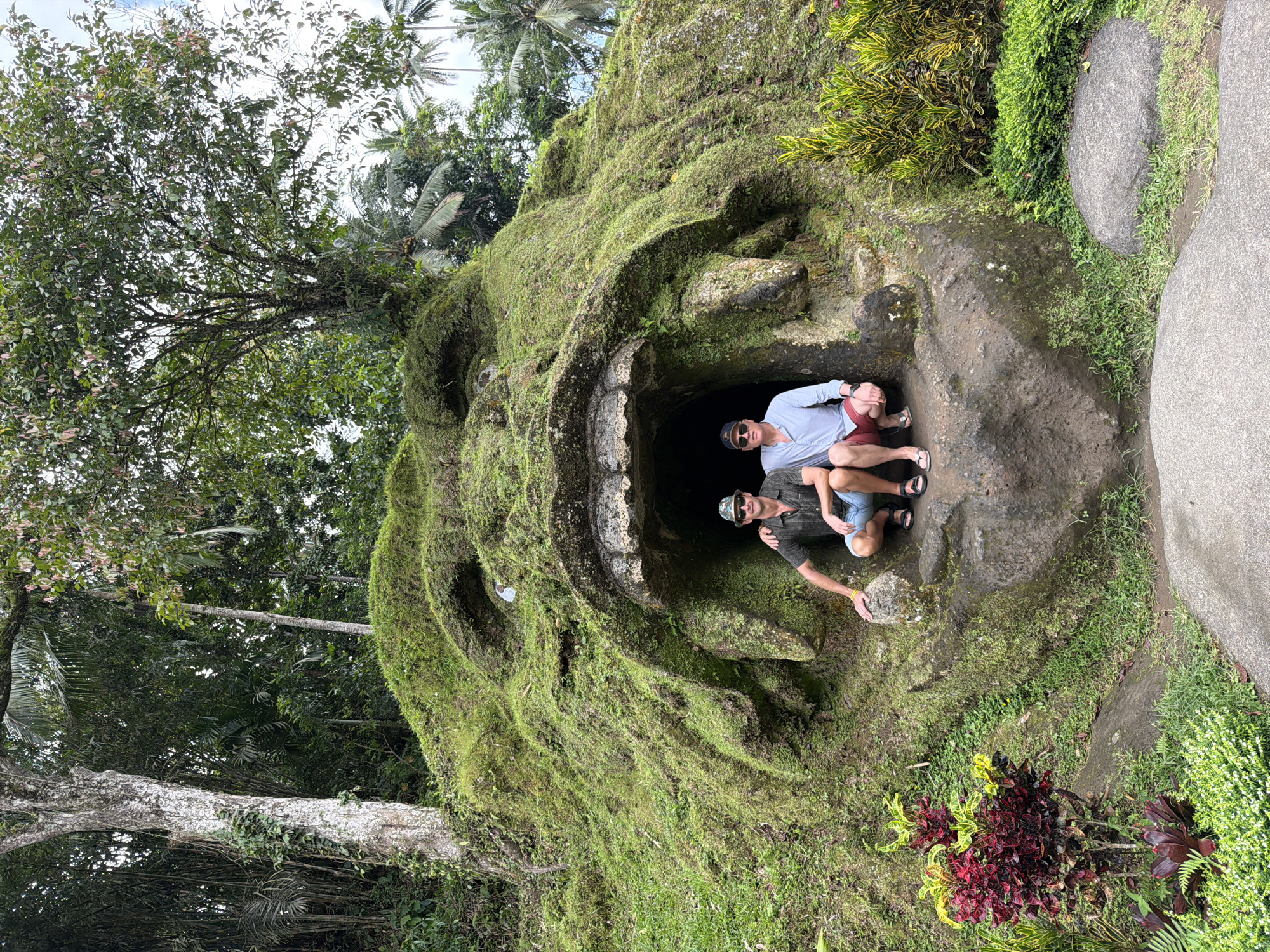 Two men are positioned inside the opening of a large, moss-covered stone structure resembling a monstrous face, likely a cultural or tourist site. The first man, closest to the foreground, wears a dark green shirt, blue shorts, sunglasses, and a hat. He has his arm extended, touching the rock formation. The second man, behind the first, wears a light blue shirt, red shorts, and sunglasses. They are both squatting inside the dark opening. The surrounding area is lush with dense tropical vegetation, including trees, plants, and grass. The image is taken at Goa Gajah Temple in Tegal Suci, Indonesia.