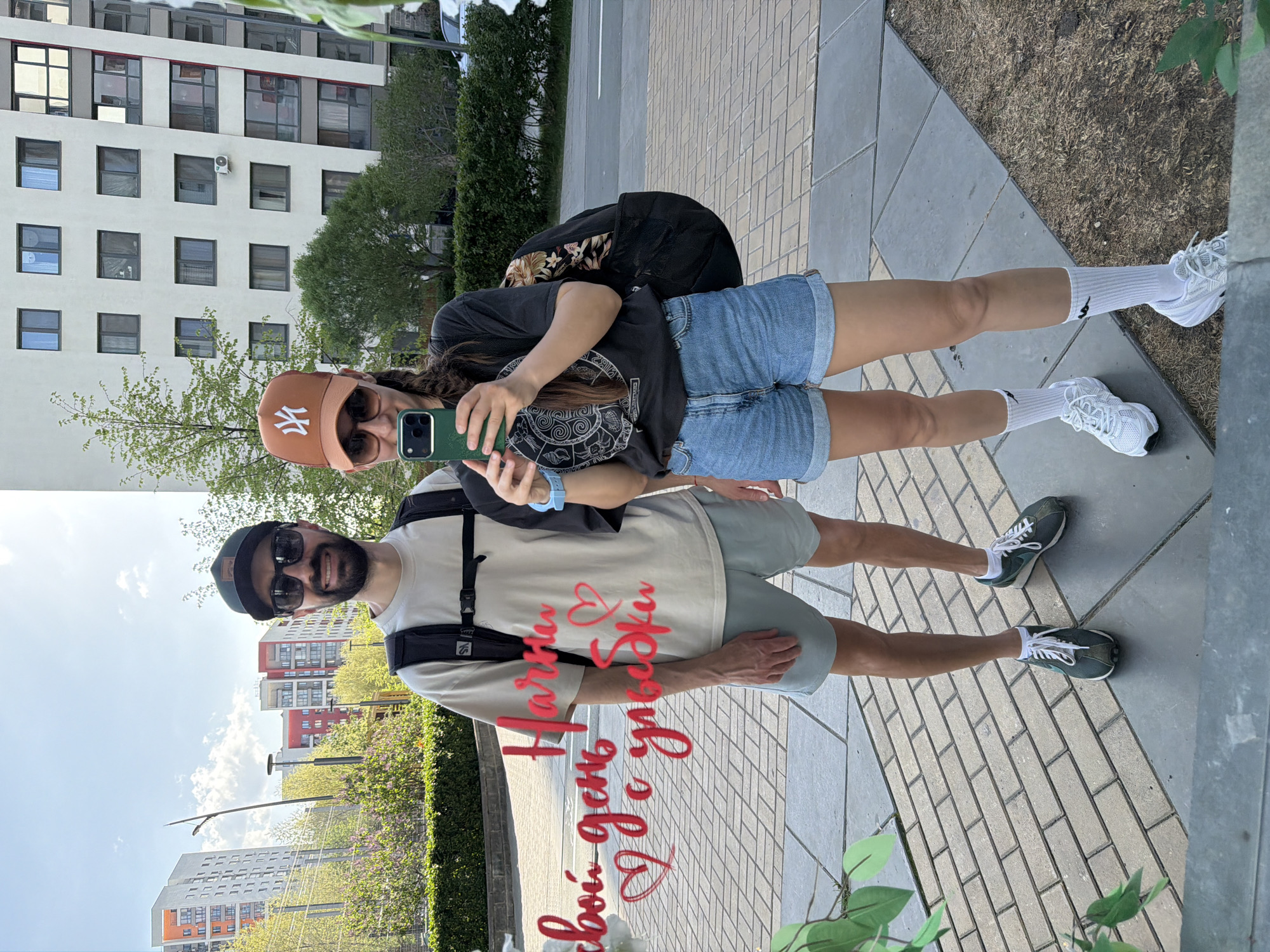 Two individuals are reflected in a flat, likely polished, surface. The person on the right is a woman wearing a black t-shirt, blue jean shorts, white socks, and white sneakers. She has a tan New York Yankees baseball cap and sunglasses. She holds a green smartphone, seemingly taking a photo or video. Behind her is a dark colored bag with a floral design. The person on the left is a man with a beard, wearing a light beige t-shirt, green shorts, and green sneakers. He is wearing a black baseball cap and sunglasses and has a black backpack. There is a partially visible red colored design overlay on his t-shirt. The reflection shows a city background with tall buildings, green trees, and blue sky. Based on the signs and building architecture, the location is Tyumen, Russia. The surface reflecting the image is likely a sign or art installation in an outdoor public area.