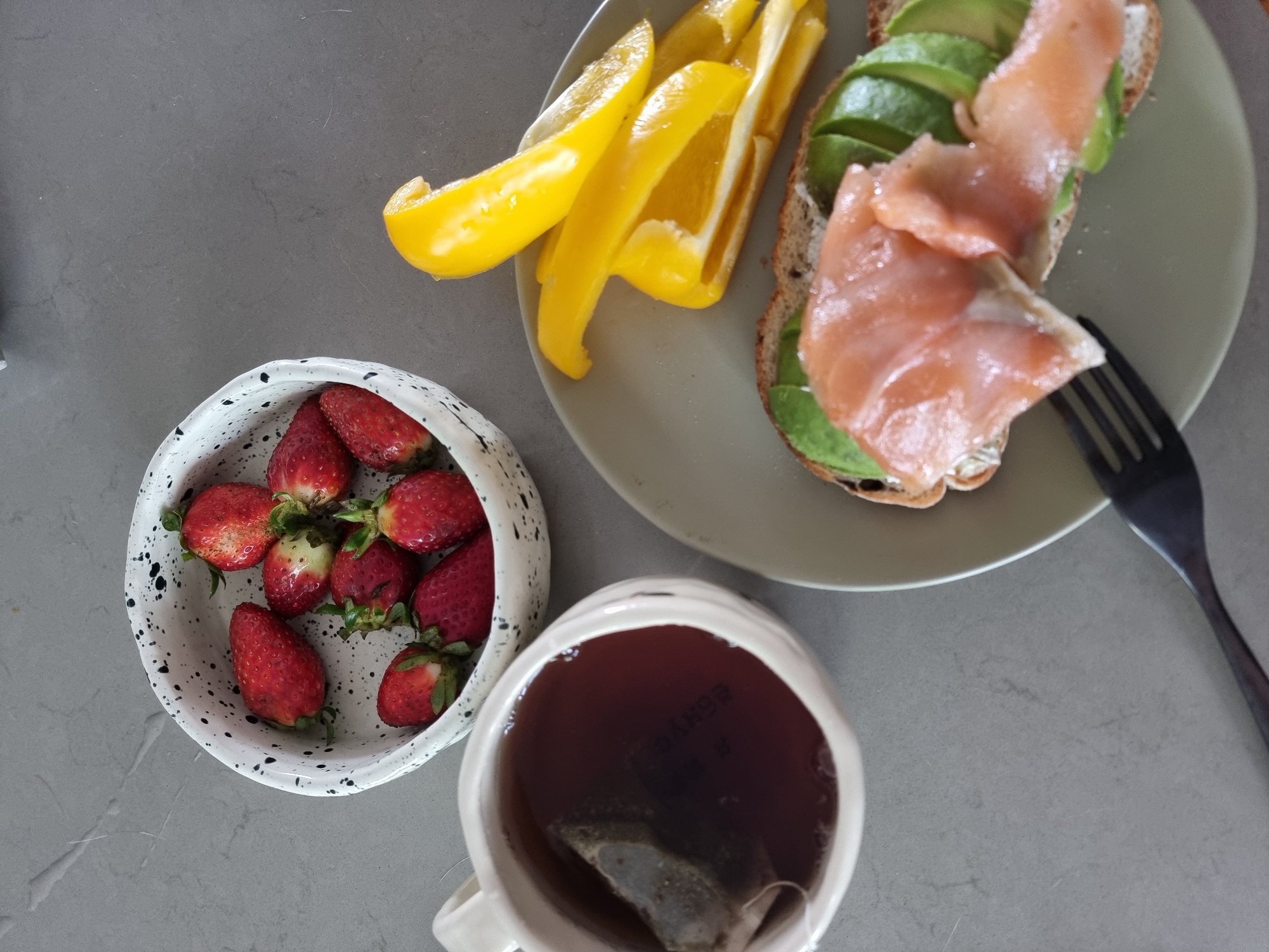 The image depicts a breakfast scene on a grey countertop. A small white bowl with black speckled pattern contains several red strawberries. A green plate holds sliced yellow bell peppers and a piece of toast topped with sliced avocado and salmon. A black fork rests on the plate near the toast. A white mug filled with dark tea, a tea bag visible inside, sits below the plate. The image appears to be taken indoors in a residential setting. While the location is Ramat Gan, Israel, there are no elements in the image to confirm this.
