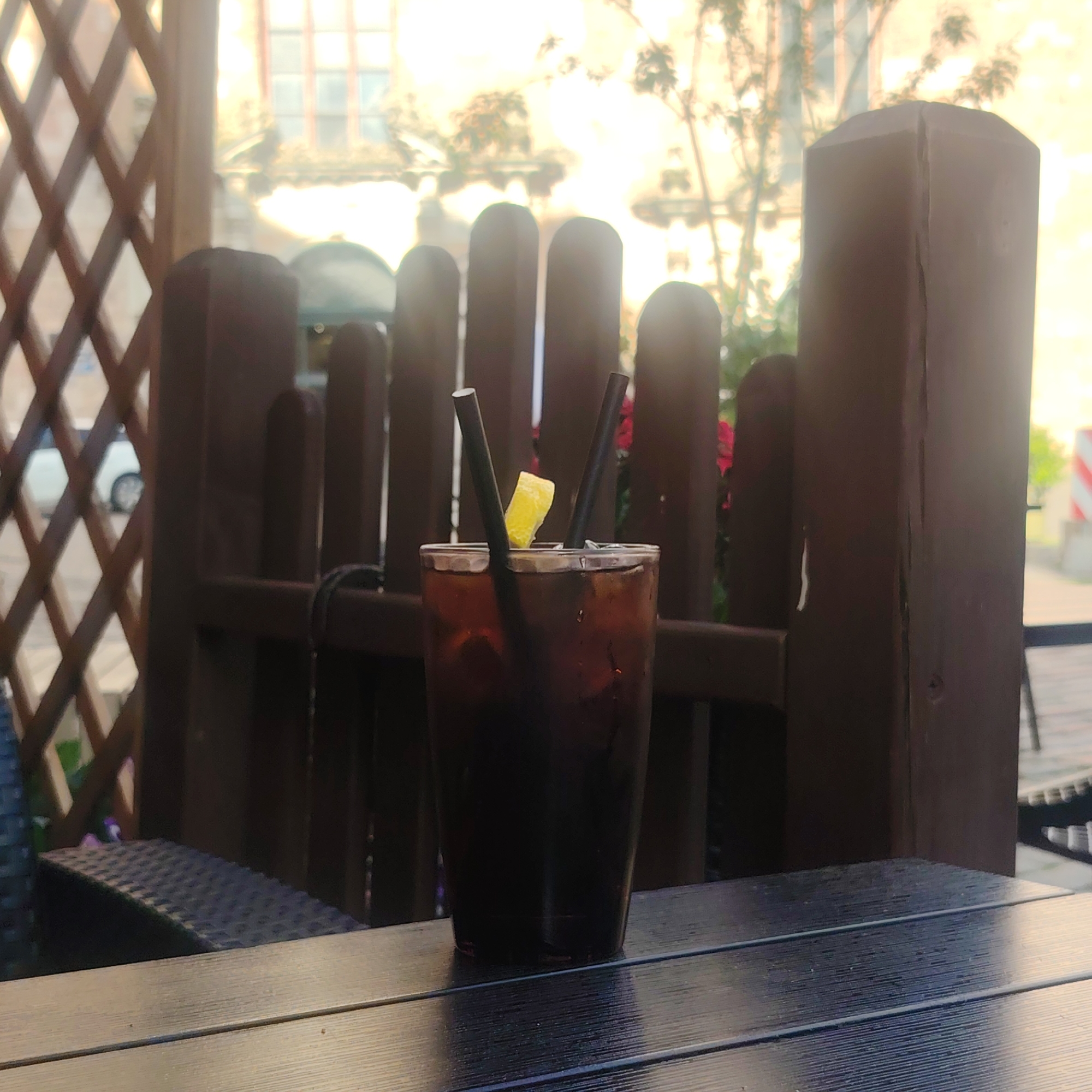 A dark brown, iced beverage in a tall glass is positioned on a dark wooden table. The drink is garnished with a slice of lemon and two black straws. The glass is positioned in front of a dark wooden fence with vertical slats and a diamond-patterned lattice screen to the left. Beyond the fence, trees and buildings are visible, bathed in bright, soft sunlight, suggesting an outdoor setting. The overall scene has a relaxed, possibly cafe or restaurant atmosphere. The image does not contain enough information to determine a specific location within Riga, Latvia.