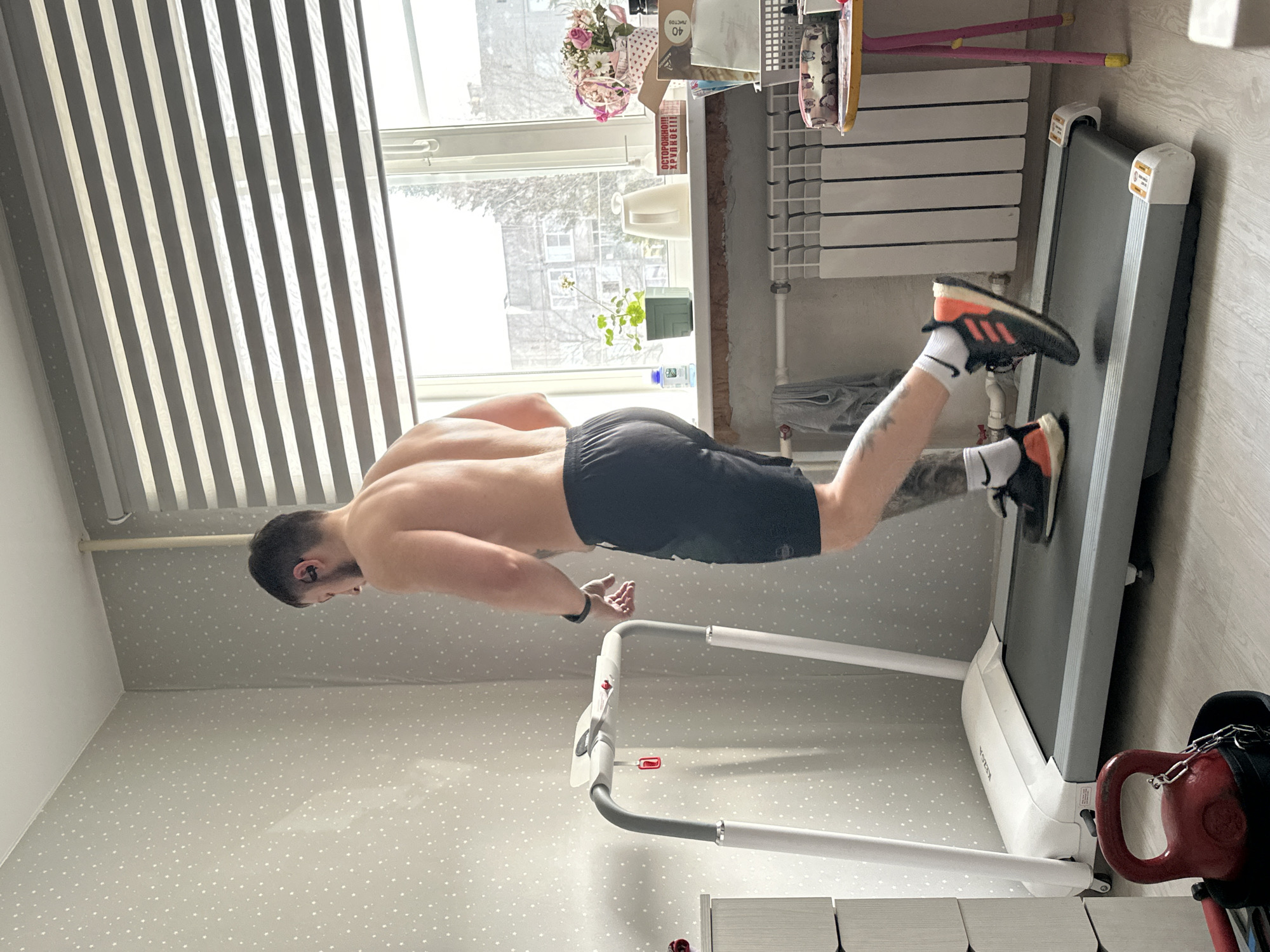 A man is performing a push-up with his feet on a treadmill. The man is shirtless and wearing black shorts, socks, and running shoes. The treadmill is white and gray. The room has a wallpaper with white polka dots. There is a window with blinds. A kettlebell is visible near the treadmill. The city and specific location are Unknown.