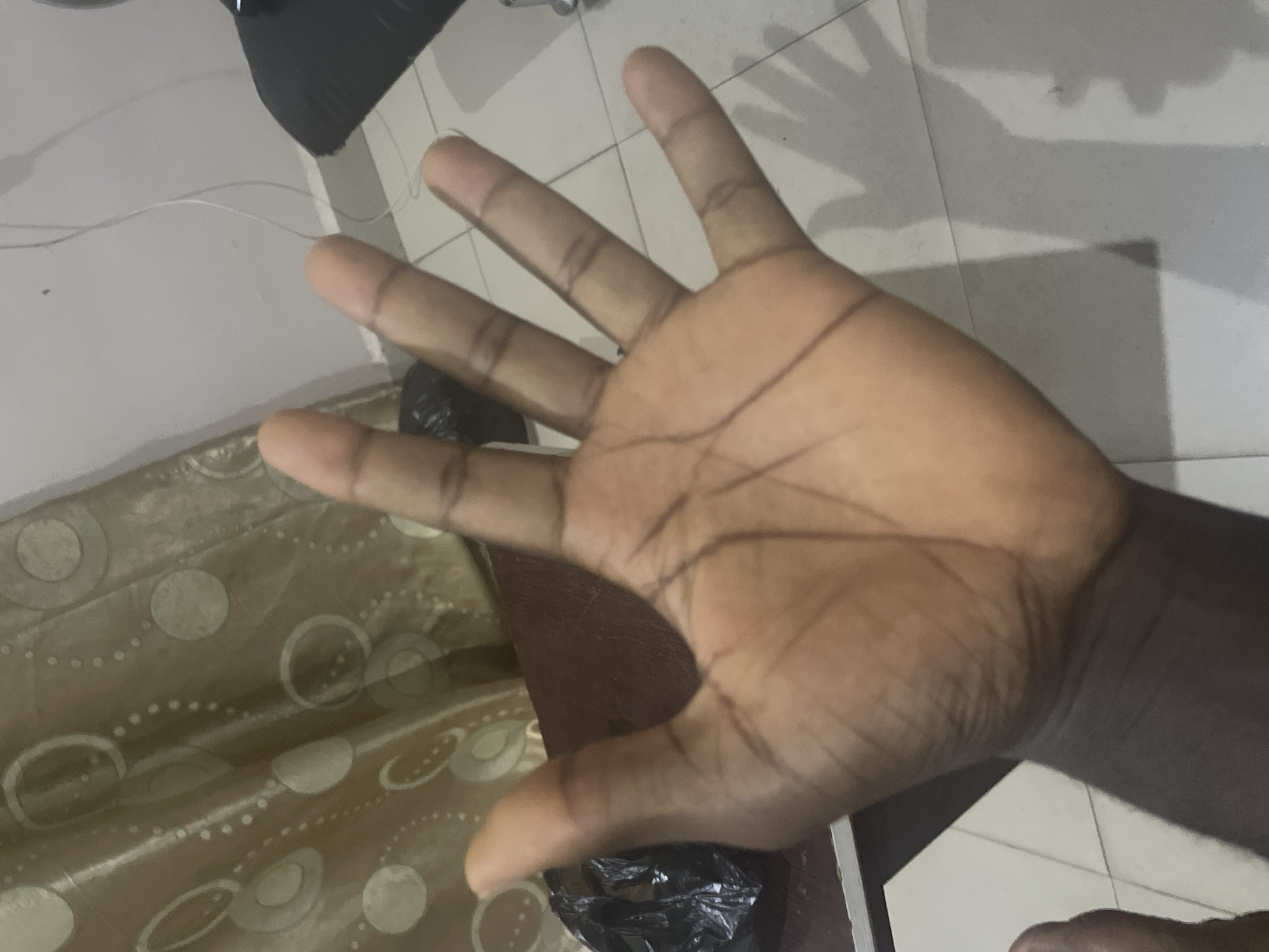 In Jalingo, Nigeria, a close-up shot focuses on the palm of an open left hand, fingers splayed. The hand is positioned in the foreground, obscuring much of the background. A crumpled black plastic bag rests near the base of the thumb, partially visible. Behind the hand, a patterned curtain with circular motifs hangs, with its folds creating a textured backdrop. The floor appears to be tiled, with a subtle shadow of the hand cast upon it. The lighting suggests an indoor setting.
