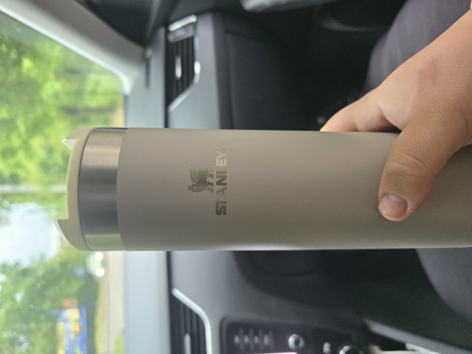 A tan Stanley brand tumbler is being held in a right hand. The scene appears to be within the interior of a car. The dashboard of the car is visible, and through the car window, there is a view of trees and a street with buildings. It is not possible to confirm that the photo was taken in the city of Pleven, Bulgaria.