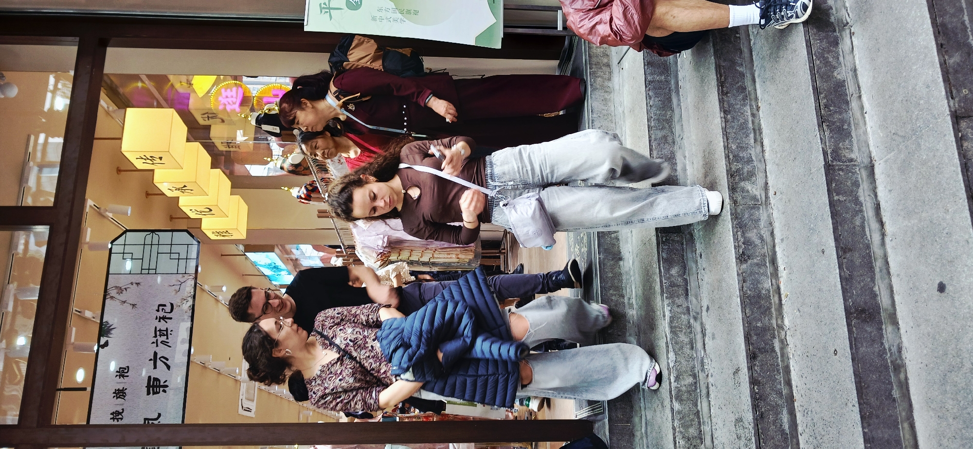 The image depicts a group of people standing on a set of concrete stairs in Yuzhong District, China. A woman with curly hair and a brown shirt is looking at her mobile phone. An Asian women in a red dress is standing behind her. A man with glasses is standing behind and to the left of the curly-haired woman. To the left of the man, is a woman wearing a floral shirt and a blue jacket. In the background are several hanging square light fixtures. Chinese characters on a vertical banner in front of a storefront are to the left. The stairs appear to be made of concrete and have dark grooves.