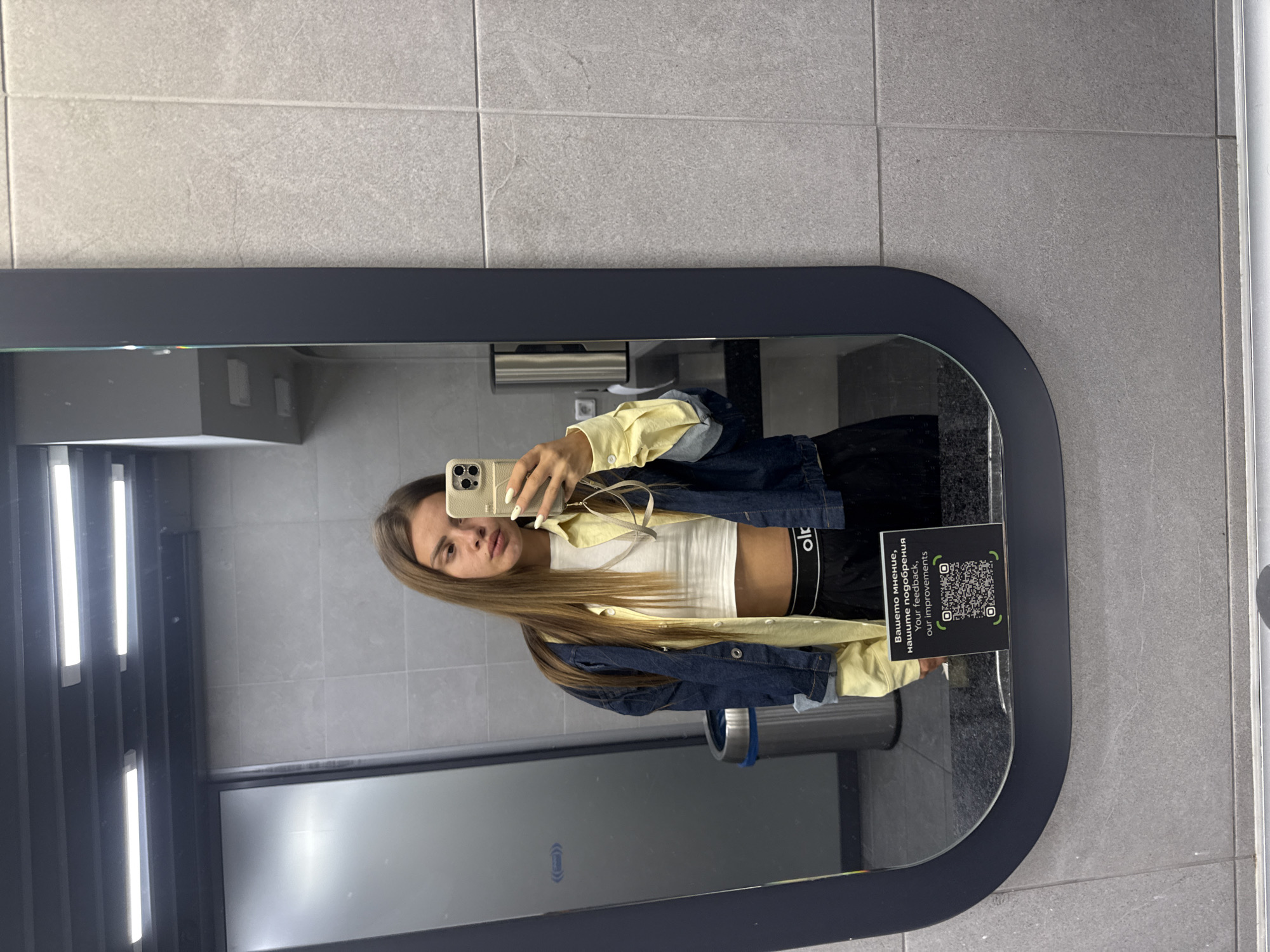A young woman with long, brown hair stands in front of a large, dark-framed mirror in what appears to be a public restroom. She is taking a selfie with a smartphone held in her right hand. She is wearing a white crop top, a yellow collared shirt worn open over the crop top, and a dark denim jacket. Her lower body is clad in black shorts with a visible white waistband.
The restroom environment features gray tiled walls. To the left of the woman, a silver trash can with a blue lid is visible. Below the mirror, a QR code is displayed on a small sign with text in both Cyrillic and English, reading "Вашето мнение, нашите подобрения / Your feedback, our improvements." The overall impression is of a modern, clean public facility. The specific location within Sofia, Bulgaria, cannot be determined from the visual information provided.