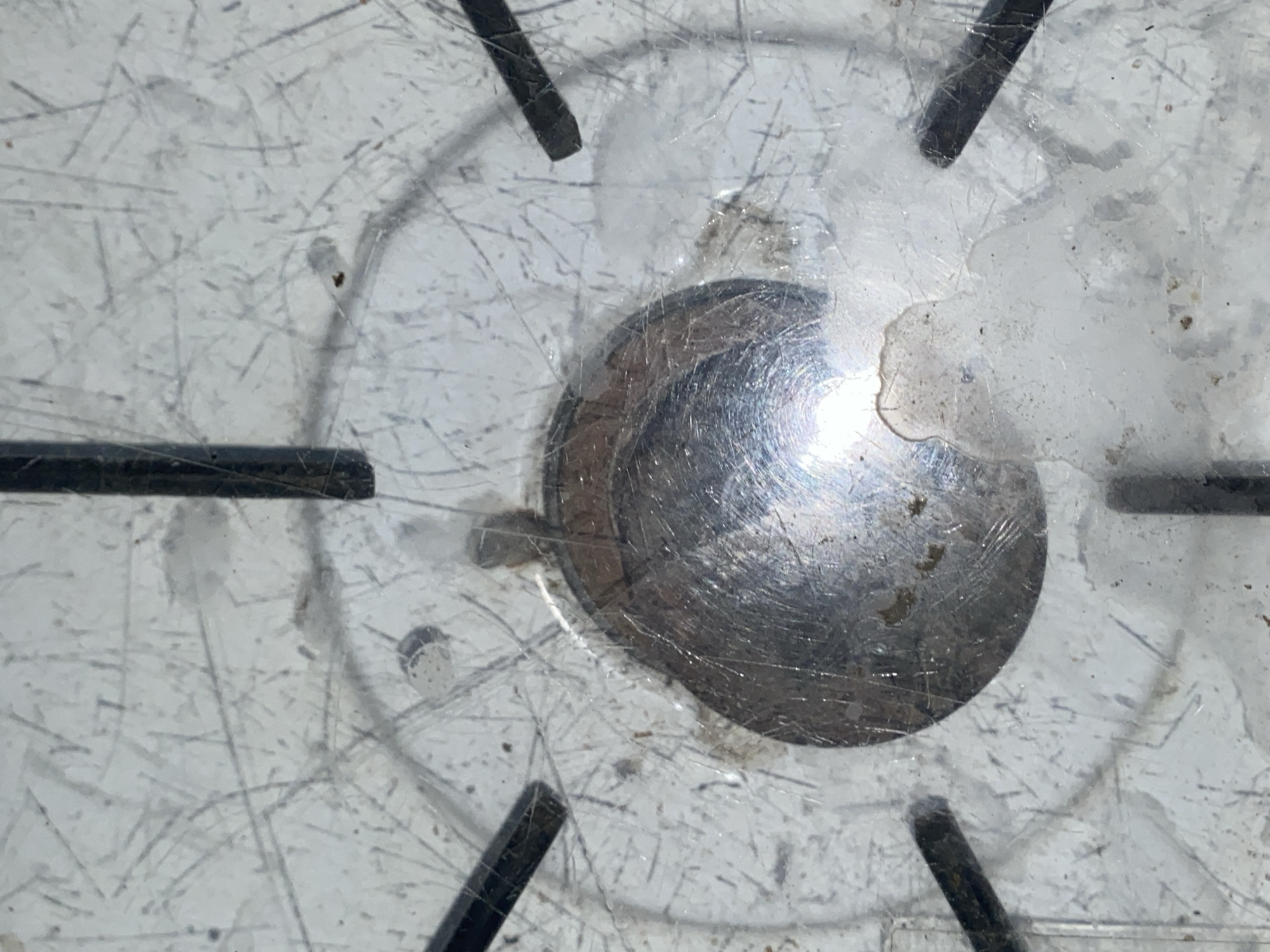 The image shows a close-up view of a scratched metal surface, possibly a stovetop, with a central drain or burner cap. The surface is a light color, possibly white or silver, and exhibits numerous fine scratches and some smudges. There are six dark, straight, rod-like objects radiating outwards from a circular area in the center. The central circular area is metallic and appears to be a raised cap or drain, with a bright reflection of light from an overhead source. Some dirt or debris is visible around the edge of the metallic central area and scattered across the scratched surface. The overall impression is of a worn and dirty surface. The image is taken from a top-down perspective.