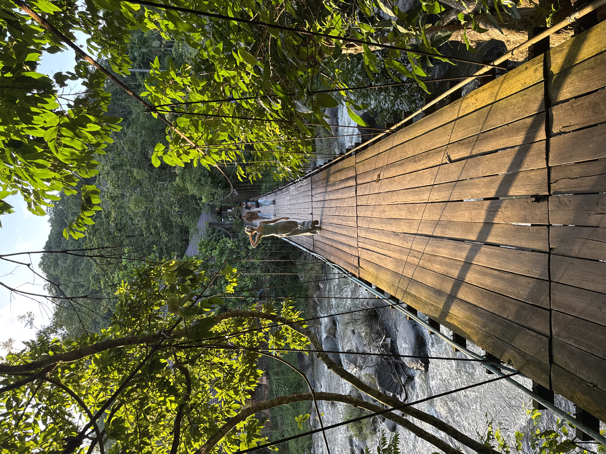 A person is lying down on a wooden suspension bridge, basking in the sun. The bridge spans over a rocky river amidst a lush green forest. Sunlight filters through the dense foliage, casting shadows across the wooden planks. The scene suggests a serene and relaxed atmosphere, likely during daytime with clear weather. The person's posture and the peaceful setting evoke a sense of tranquility and enjoyment of nature. The location, Ranong, Thailand, hints at a tropical environment.