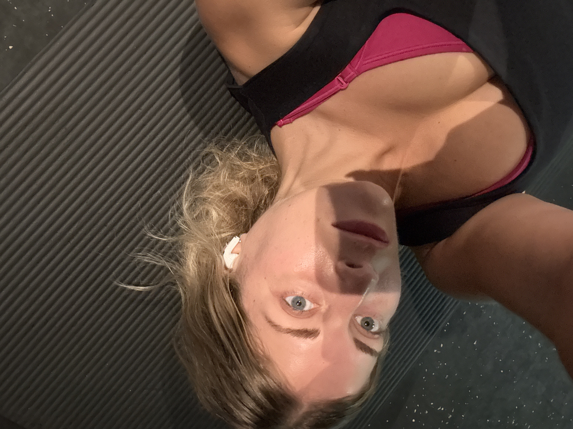 A person with blonde hair and blue eyes is lying on a black yoga mat. They are wearing a black tank top and a pink sports bra. The person is looking up at the camera, and their face is illuminated by a light source from the side, creating shadows. In the background, there are some white specks on a dark surface. The environment appears to be indoors, possibly a gym or studio. There is no indication of the city of Paris, France in the image.