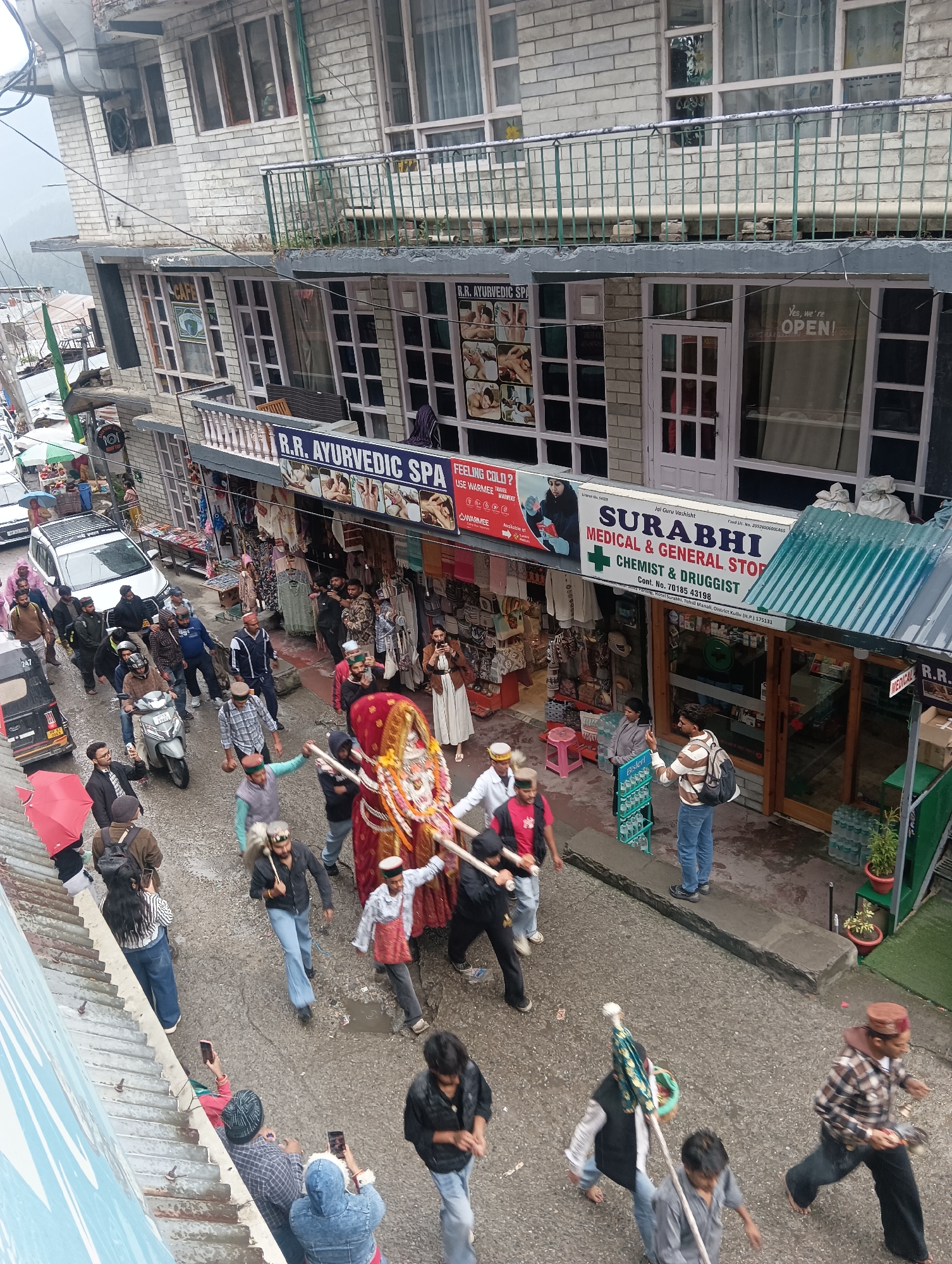 This image captures a lively street scene in Vashisht, India, likely during a procession or festival. The view is from above, looking down onto a narrow, wet street lined with shops. In the foreground, several people are walking, some carrying what appears to be a decorated palanquin or float adorned with red and gold fabric. One person in the procession wears a traditional Himachali cap.
The street is crowded with people, many dressed in casual clothing, and some are holding umbrellas, suggesting it might be raining or has recently rained. A scooter is being ridden by one man, and an auto-rickshaw is parked on the left side of the street. Shops with various signs are visible along the street, including "R.R. Ayurvedic Spa," "Cafe," and "Surabhi Medical & General Store." The buildings are multi-story, with balconies and windows overlooking the street. The overall atmosphere appears to be bustling and festive, with a mix of commercial activity and cultural celebration.
