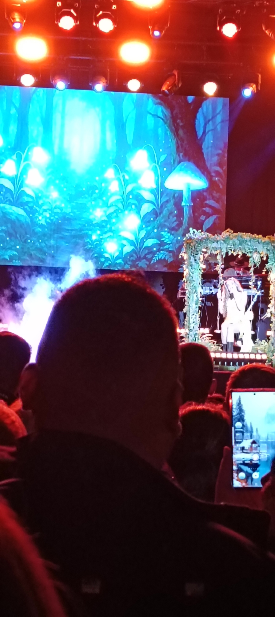 The image shows a live performance. In the foreground, an audience member's head partially obscures the view. To the right of that head, another audience member is holding a smartphone, appearing to record or photograph the performance. On stage in the background, a performer is seated at a keyboard, within a structure decorated with foliage. Behind the performer, a large screen displays a forest scene with glowing plants and mushrooms. Above the screen, stage lights are visible. The location is likely a performance venue, possibly in Cerdanyola del Vallès, Spain.