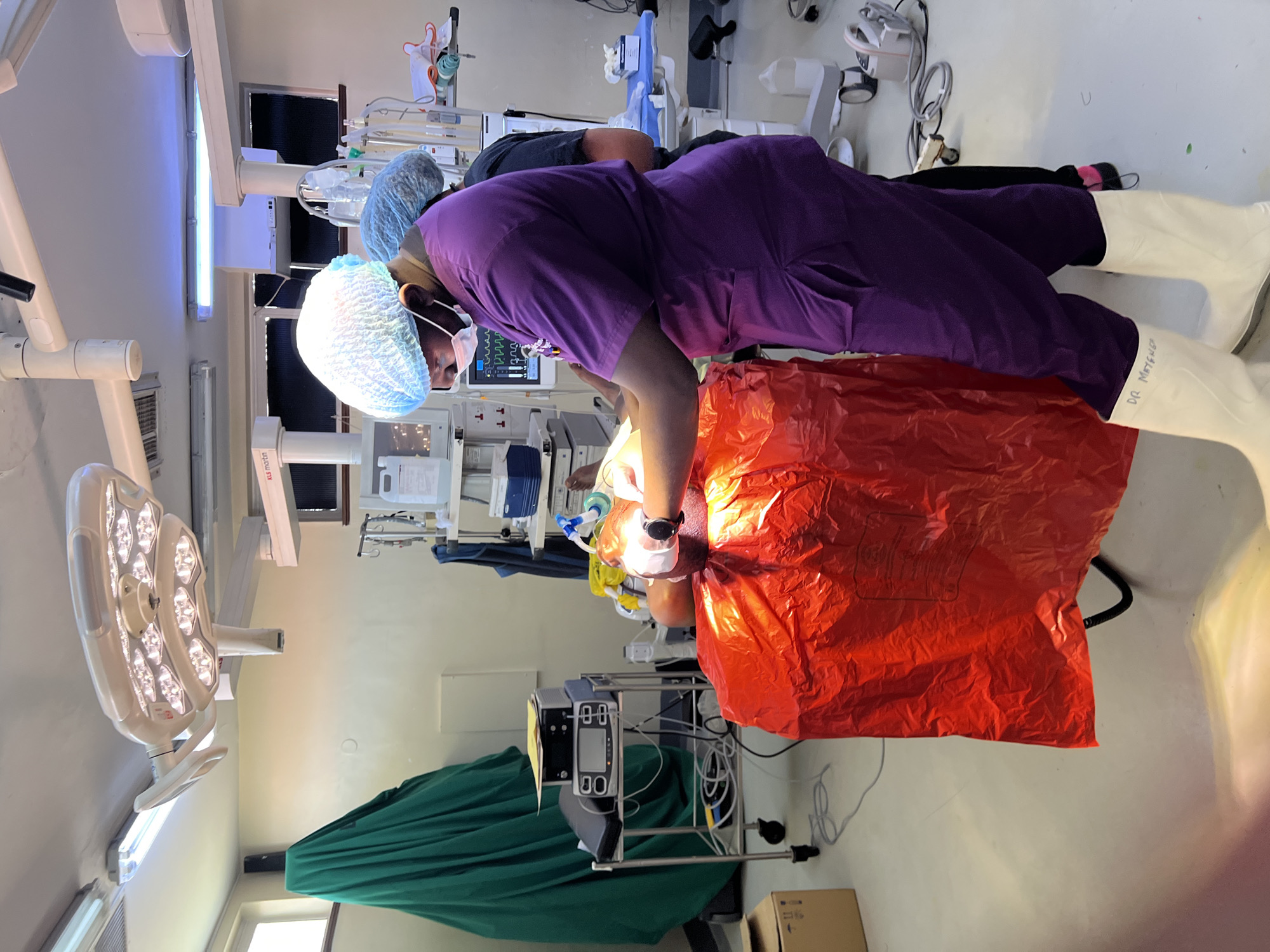A surgery is in progress in a medical operating room, possibly in Nairobi, Kenya, given the inscription "DR. Ndufungu" on a person's white overshoe. An individual in purple scrubs and a surgical cap is leaning over a patient covered with an orange surgical drape. Monitoring equipment, including screens, are visible in the background, along with other medical apparatus. A surgical light fixture is mounted above the scene, and a green drape is present in the lower left corner.