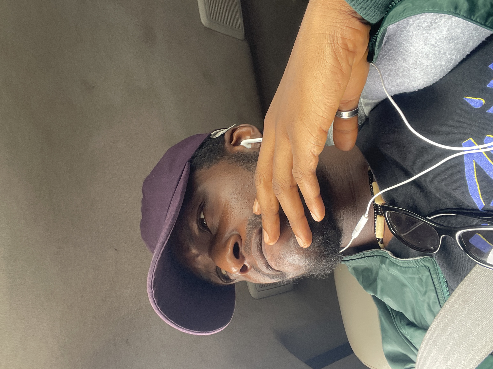 A man is in a car in Sobon-Gida, Nigeria. He is wearing a purple baseball cap, a dark t-shirt with a pattern, and a green jacket. He has earbuds in and sunglasses next to him. He is wearing a seatbelt and a ring on his finger. His hand is partially covering his face. The car interior is visible.