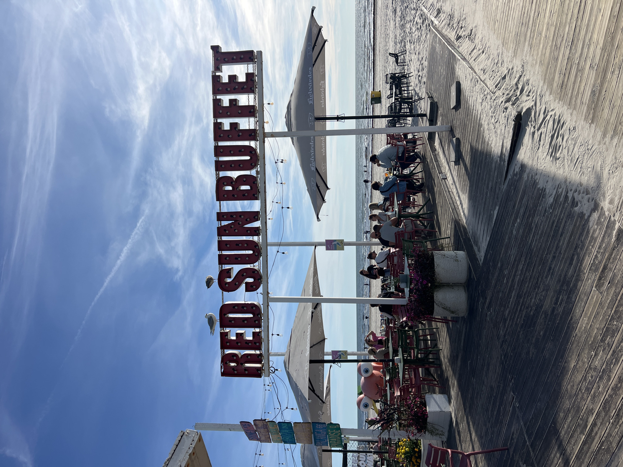 An outdoor dining area of "Red Sun Buffet" is situated on a wooden boardwalk adjacent to a sandy beach. The restaurant features numerous tables and chairs, some with umbrellas, and is decorated with a large pink inflatable frog. In the background, the sea is visible with several people on the beach and in the water. Two seagulls are perched on the restaurant's signage. The scene suggests a pleasant, sunny day. The location is the beach in Liepaja, Latvia.