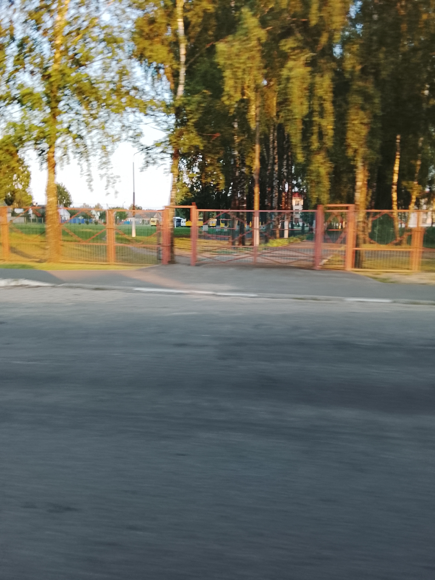The image captures a fence in front of a park or schoolyard in Zhlobin, Belarus. The fence is made of orange metal bars and appears to be a double gate. Behind the fence, there are trees with green and yellow leaves, indicating it is likely autumn. In the background, there are buildings and a street. The foreground shows a blurred asphalt road with a white line marking the edge. The image is taken from a moving vehicle, resulting in motion blur.