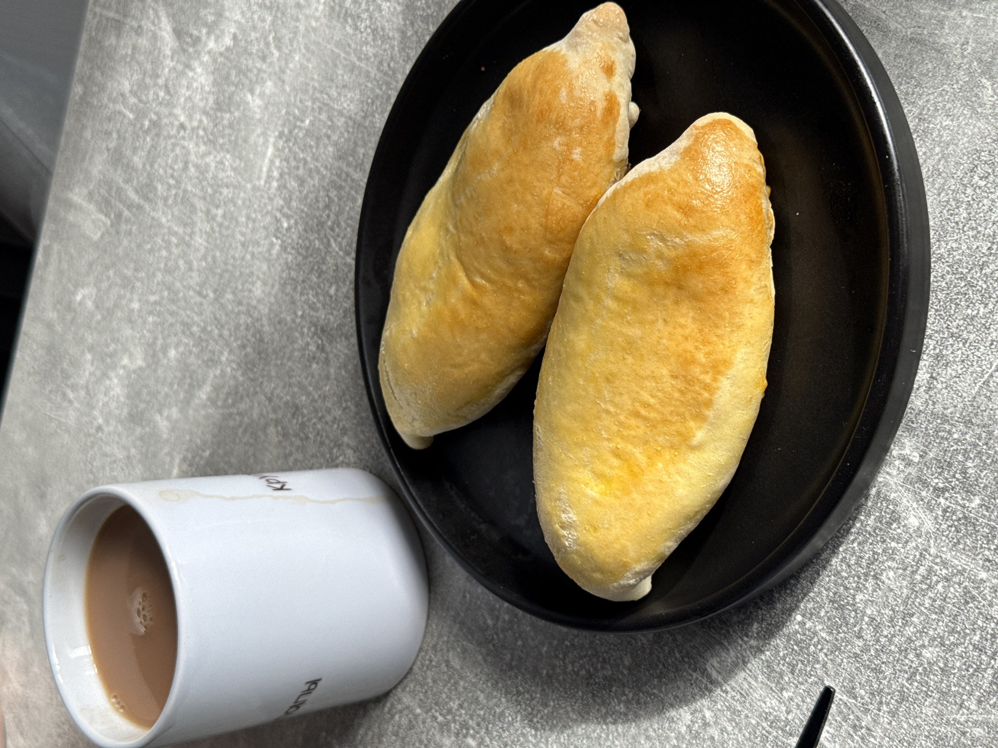 An overhead, medium shot shows two golden-brown, oblong pastries on a black plate. The pastries are positioned slightly angled, with the bottom one closer to the camera and the top one slightly behind and to the right. They appear to be baked goods, possibly filled, with a slightly uneven, toasted surface. To the left of the plate, a white ceramic mug is visible, filled with a light brown liquid, likely coffee with milk. A thin, light-colored drip runs down the side of the mug. The items are placed on a textured, gray surface that resembles a concrete countertop. There are no people visible in the frame. The lighting suggests an indoor setting, with a shadow cast from the mug onto the surface to its left. There is no information available about the location in Lyubertsy, Russia.