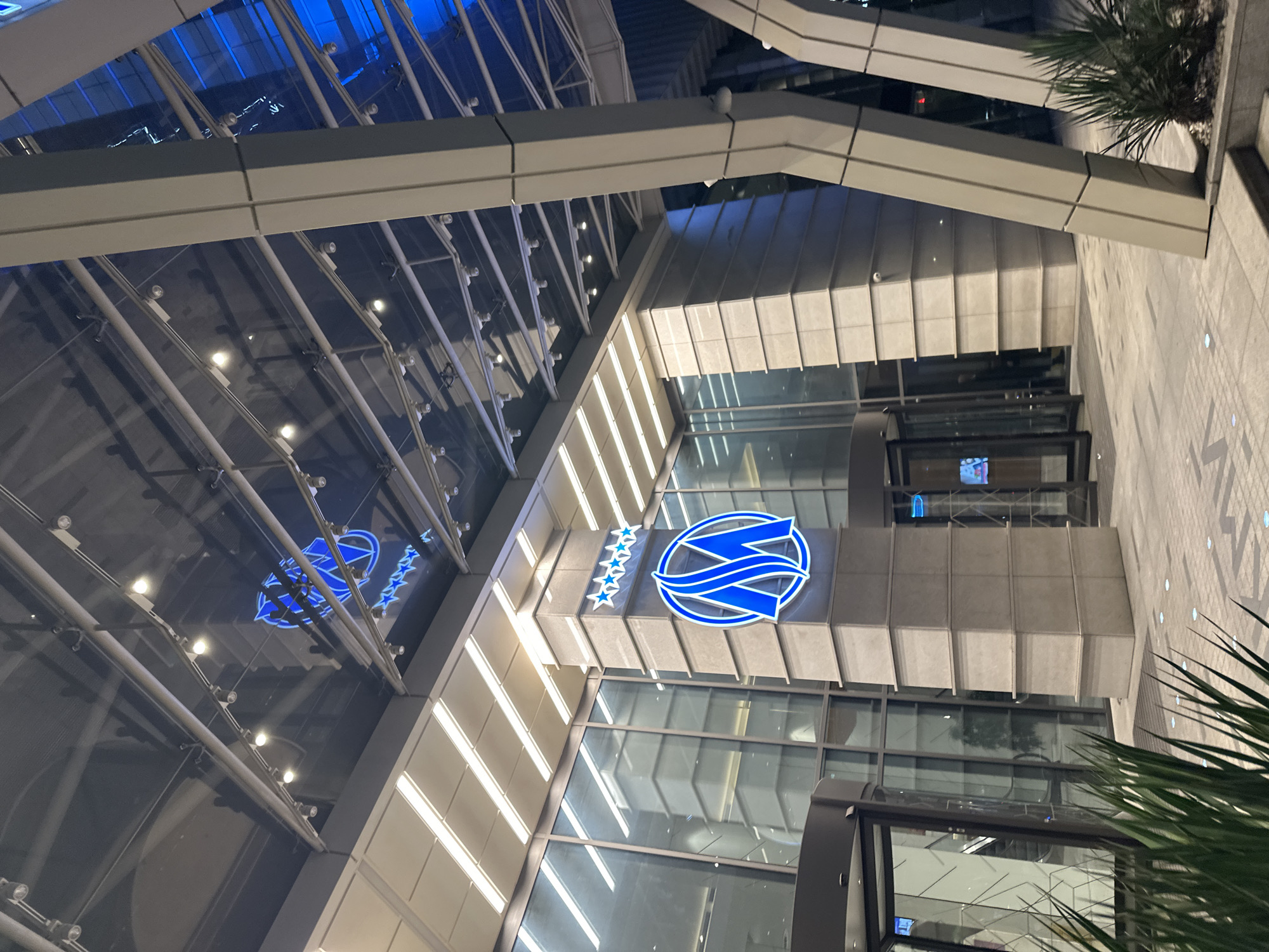 An exterior shot captures the entrance of a modern building, possibly a hotel or office building, in an urban setting at dusk or night. The architecture features a large glass ceiling supported by a network of metal beams, reflecting the lights from within and the sky above. The building's facade is a mix of glass, concrete, and metal. A prominent column near the entrance displays a blue neon logo with five stars above it. Two revolving glass doors are visible, one partially open. In the foreground, green plants add a touch of nature to the urban landscape. The ground is paved with concrete tiles. The lighting is predominantly blue and white, creating a sleek and contemporary ambiance.