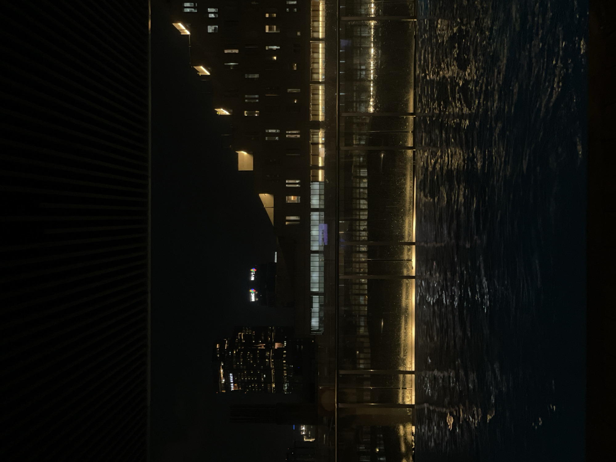This is a nighttime photograph taken from a high angle, likely from a high-rise building in Central Jakarta, Indonesia. The main elements visible are buildings with lit windows, a waterfall feature, and an apparent water surface. The buildings on the left have a dense concentration of illuminated windows, suggesting a high occupancy rate. Signage is visible, and it can be determined that one of these buildings is a 'Park Hyatt' hotel. To the right, there's a waterfall structure with vertical sections and illuminated edges, creating reflections on what seems to be a pool of water. The overall lighting is low, creating a contrast between the dark sky and the lighted buildings and waterfall. The photograph appears to be taken at a sharp angle.
