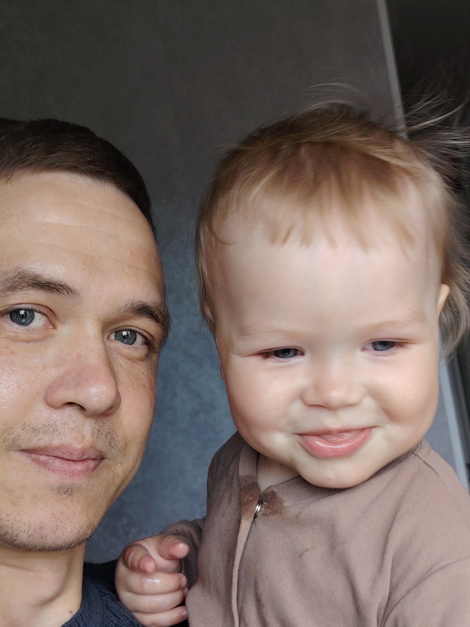 A close-up, indoor portrait features an adult man and a baby, likely taken in Chaikovsky, Russia. The man is positioned on the left side of the frame, looking directly at the viewer with a neutral to slightly pleasant expression. He has short, dark hair, blue eyes, and light stubble on his face. He is wearing a dark top, possibly a sweater.
To the right, a baby with light blonde, slightly disheveled hair is visible. The baby has light blue eyes and a joyful, playful expression, with a slight smile and the tongue peeking out. The baby is dressed in a light brown or beige long-sleeved top, possibly a hooded onesie, with a visible zipper pull and drawstring near the neck. A small, dark or wet spot is noticeable on the baby's garment below the collar. The baby's left hand is visible, with fingers curled, resting near the man's shoulder.
The background is a plain, dark grey, textured wall, indicating an indoor setting. The lighting is soft and even. The image captures a tender, familial moment. No text is visible in the media file.