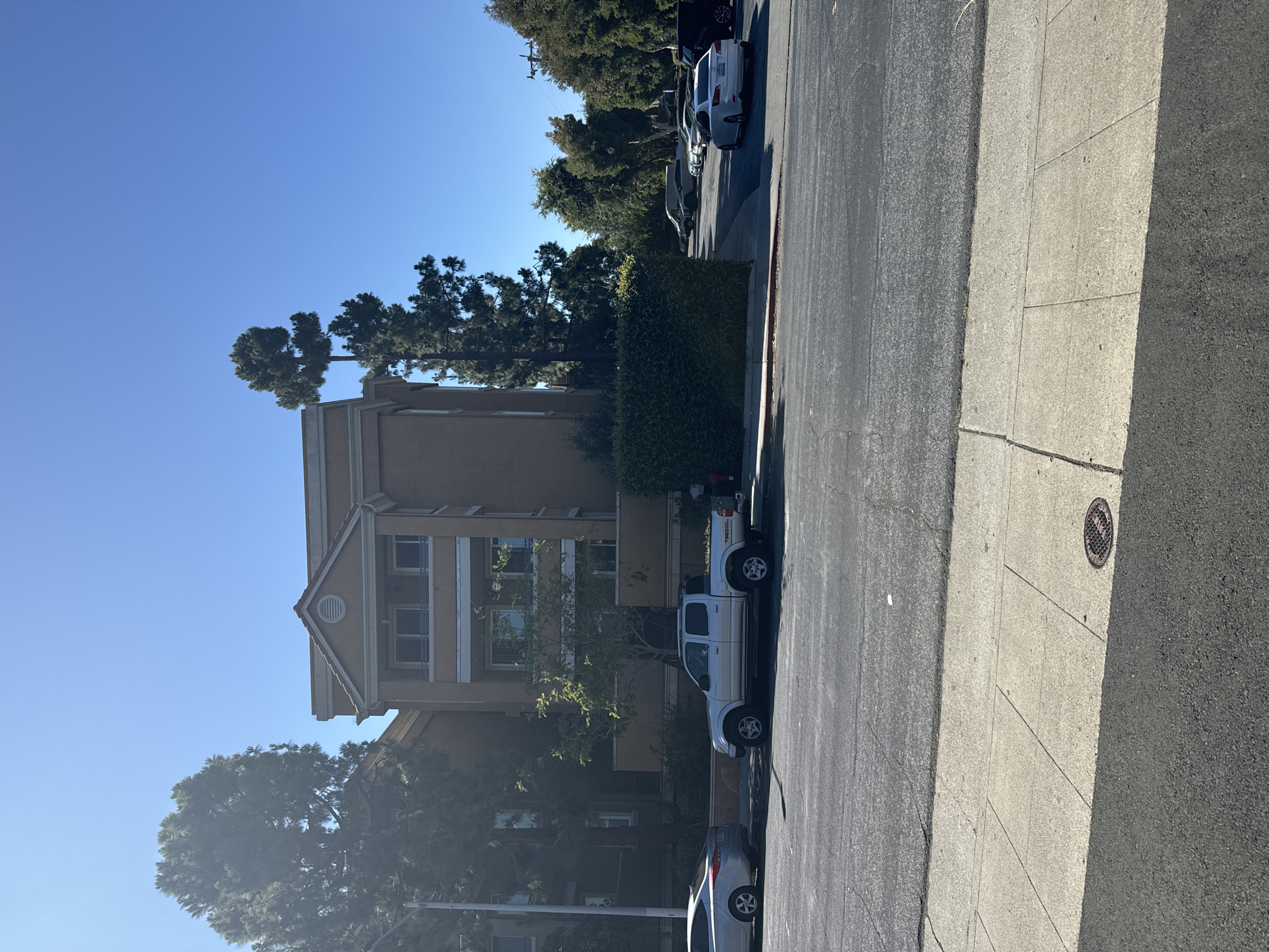 In the city of San Mateo, California, United States of America, a sunlit street scene is captured. The image features a light brown, multi-story building with a pitched roof, flanked by lush green trees and well-maintained hedges. In front of the building, a silver Toyota Tacoma pickup truck is parked. A silver sedan is parked to its left. The foreground shows a paved road with a sidewalk. The sky is clear and blue, indicating a bright day.