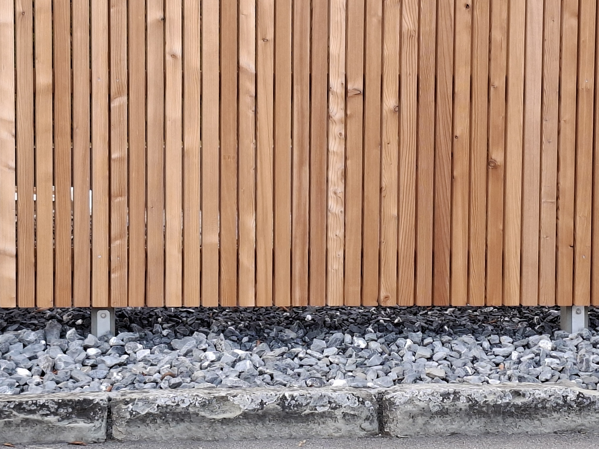 The image shows a wooden fence with vertical slats. The fence is made of light brown wood with visible grain and knots. It is supported by thin, light-colored metal posts, one of which is visible near the center of the image. Below the fence, there is a layer of dark gray gravel, topped with a layer of lighter gray, rough-textured stones. A concrete curb or edge runs along the bottom of the image, separating the gravel from what appears to be a paved surface. The scene is outdoors, with no people or animals present. The overall impression is of an exterior fence in a landscaped or utilitarian area. The location is specified as Hochdorf, Switzerland.